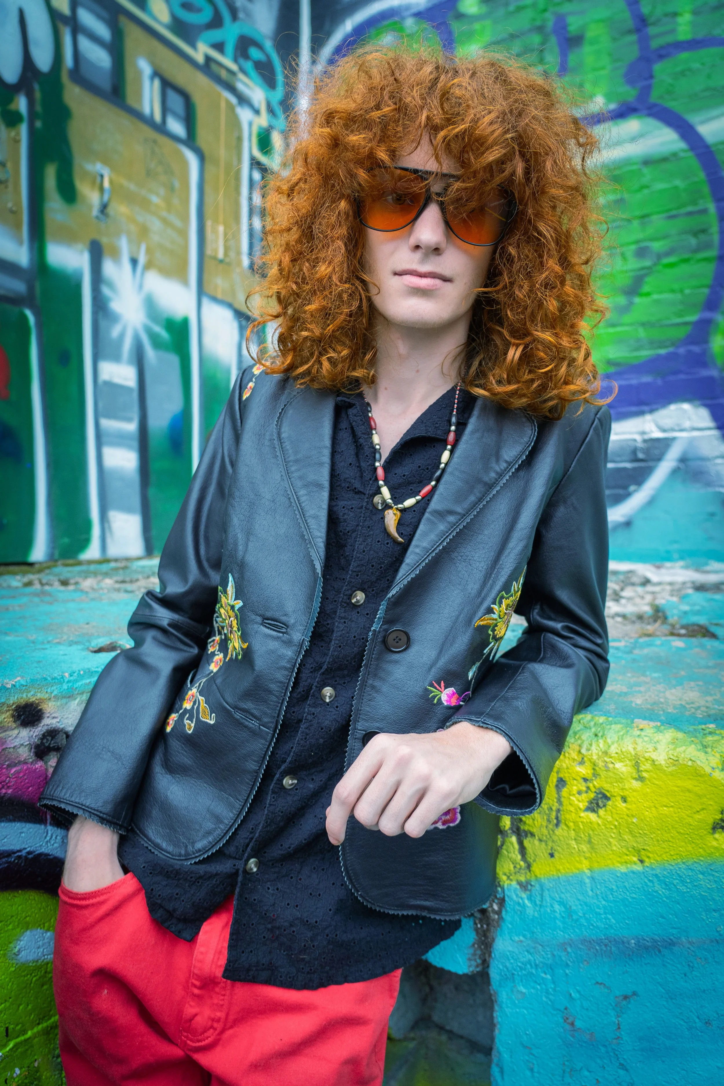 A woman with curly red hair wearing sunglasses, a black embroidered leather jacket, black blouse, with a beaded necklace, standing against a colorful graffiti wall.