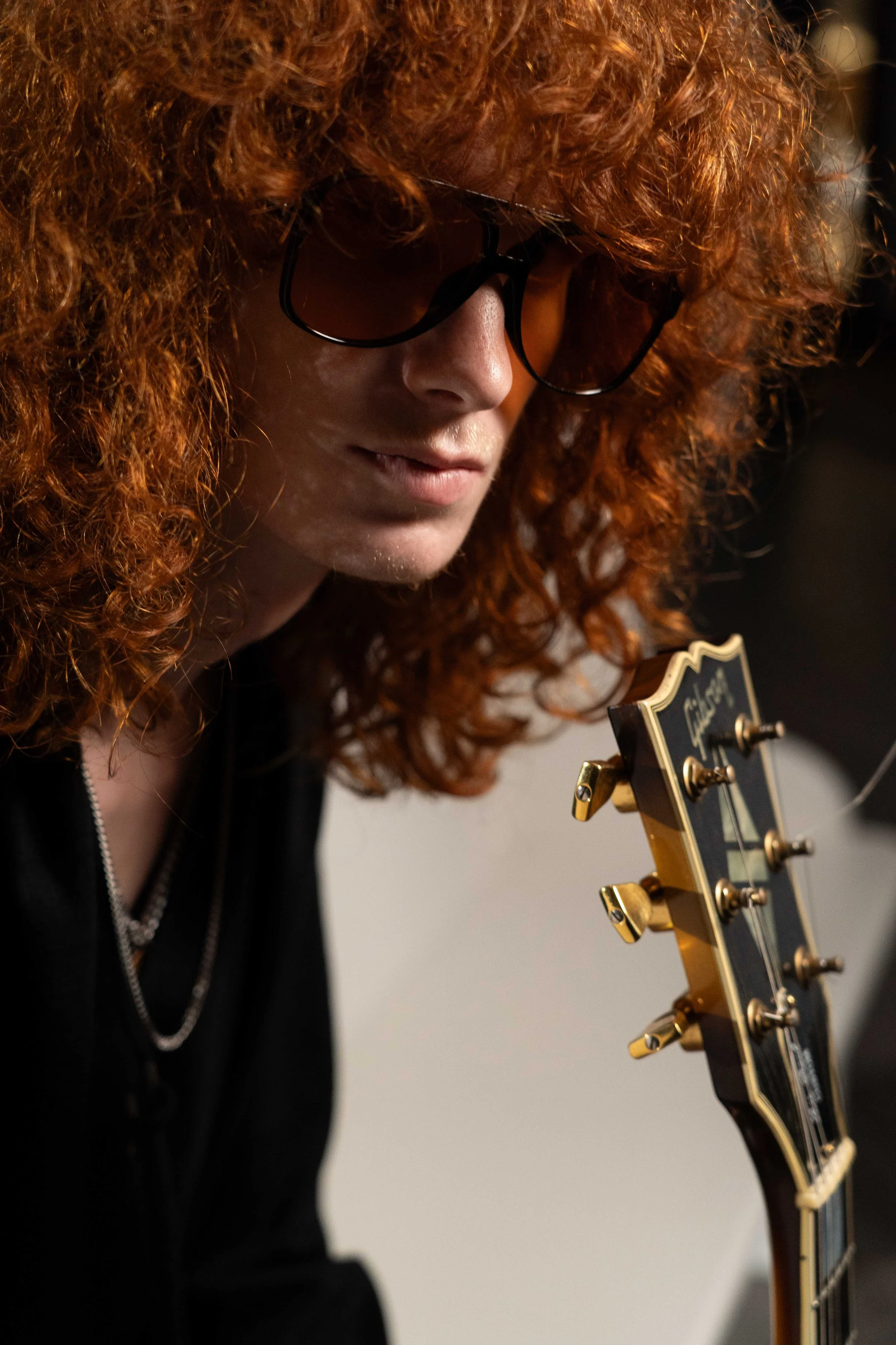 A woman with curly red hair and sunglasses looking down at a guitar.