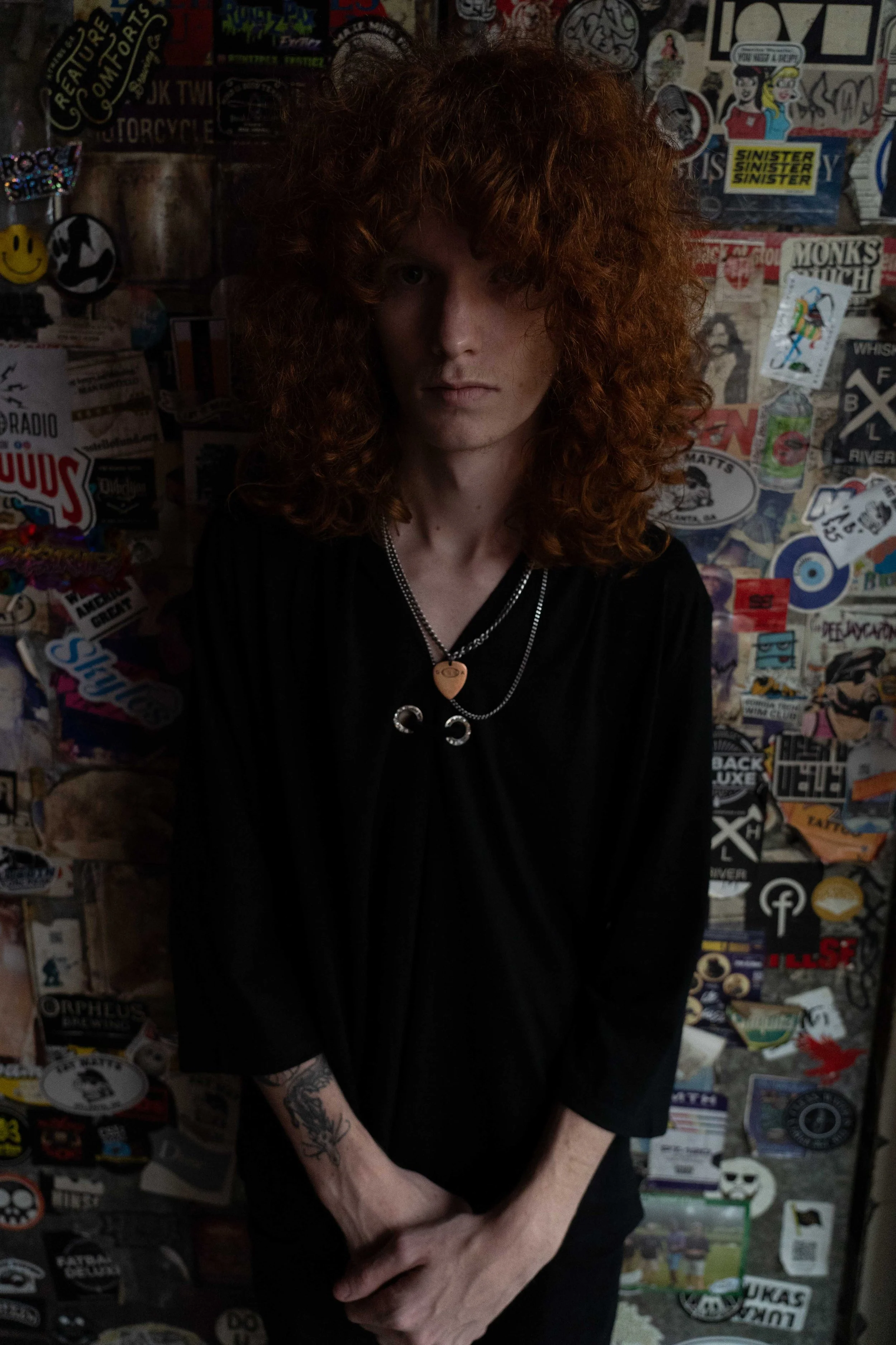 A person with long curly red hair, wearing a black shirt and layered necklaces, standing against a wall covered with colorful stickers and graffiti.