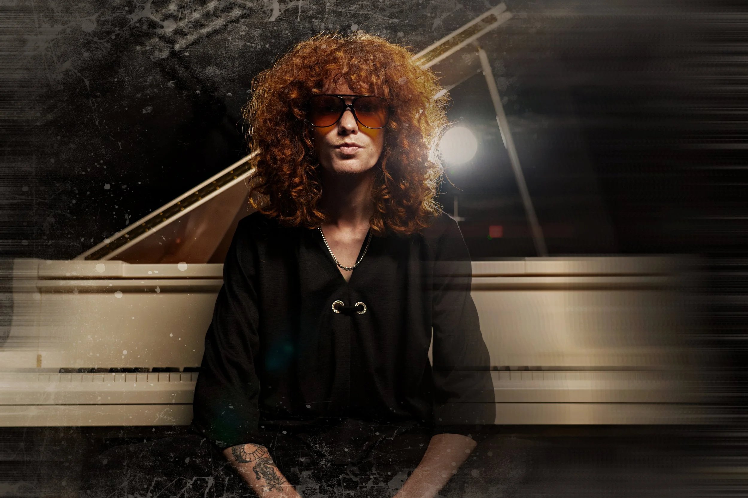 Woman with curly red hair wearing sunglasses, black top, and tattooed arm, sitting in front of a grand piano in a dimly lit setting.