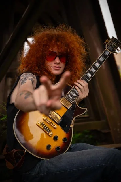 Person with curly red hair wearing red sunglasses, holding an electric guitar, making a peace sign, sitting outdoors.
