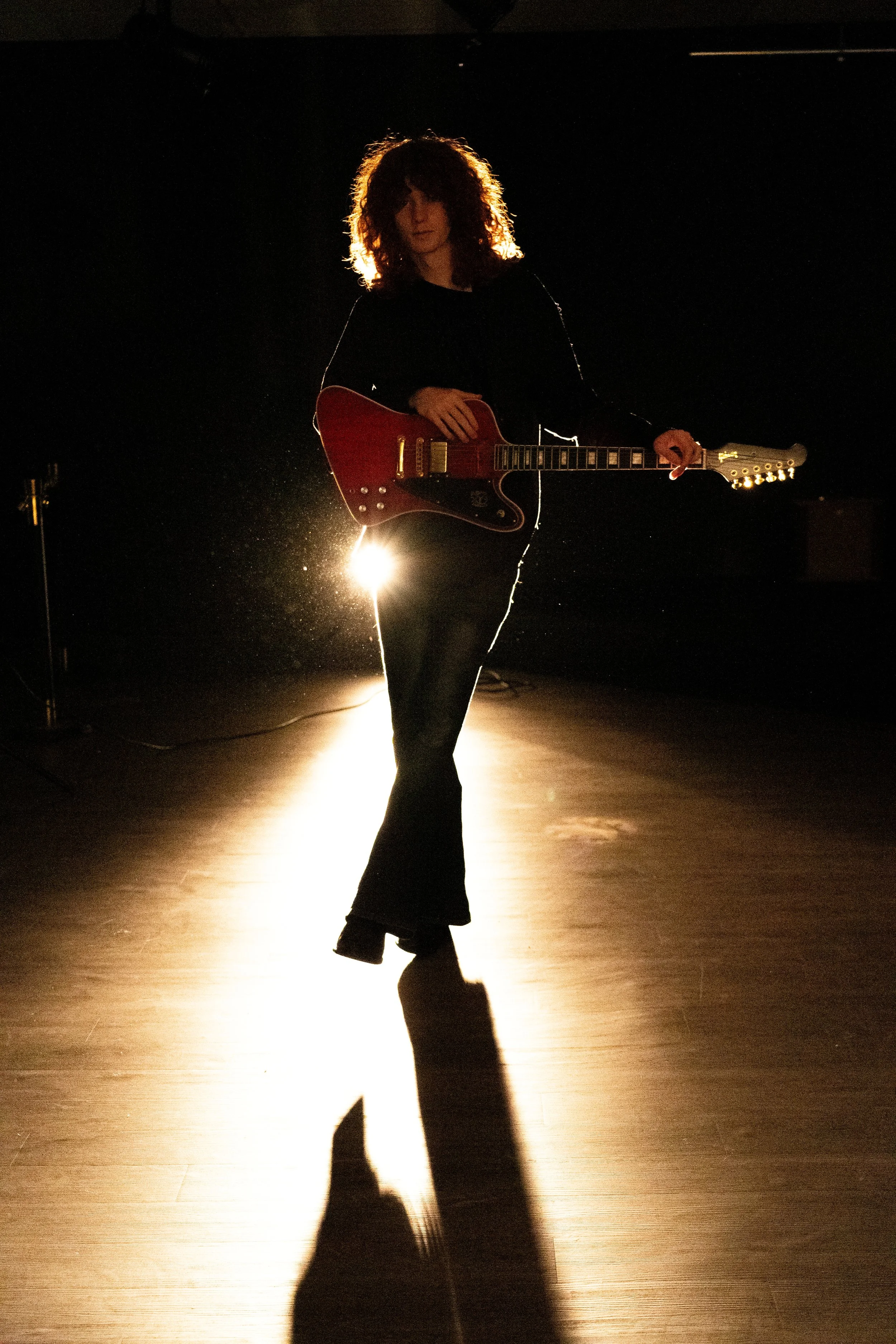 A person with curly hair holding a red electric guitar, standing on a dimly lit stage with backlighting creating a silhouette and shadow.