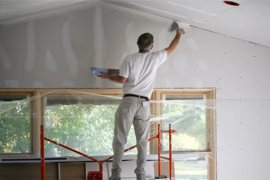 Person painting or preparing drywall on ceiling inside a room, standing on a scaffold, with large windows and natural light.