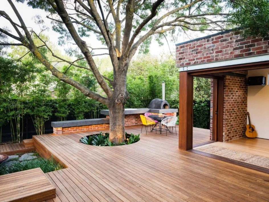 Outdoor wooden deck with a tree growing through it, surrounded by greenery and adjacent to a brick house with an open interior. There is a seating area with colorful chairs and a brick outdoor oven.