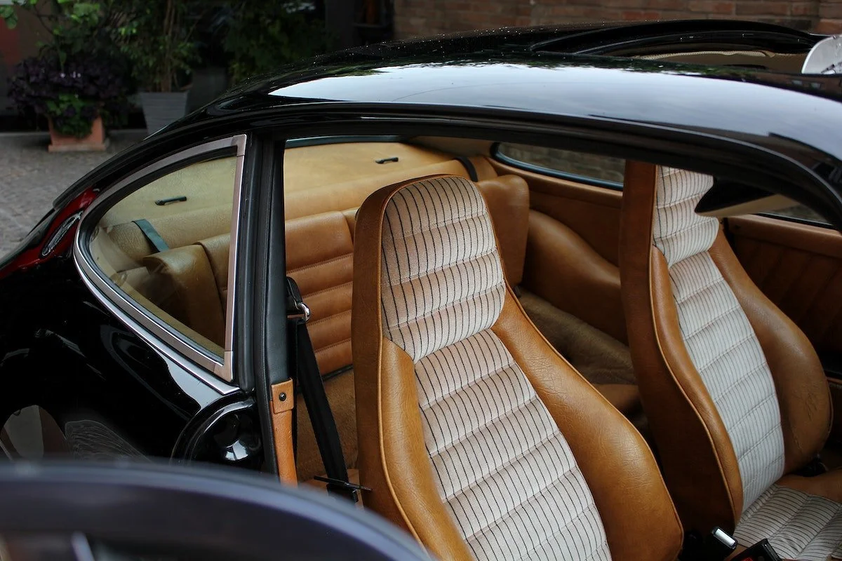 Inside of a vintage black car with tan and striped fabric seats and a view of the rear seats and side window.