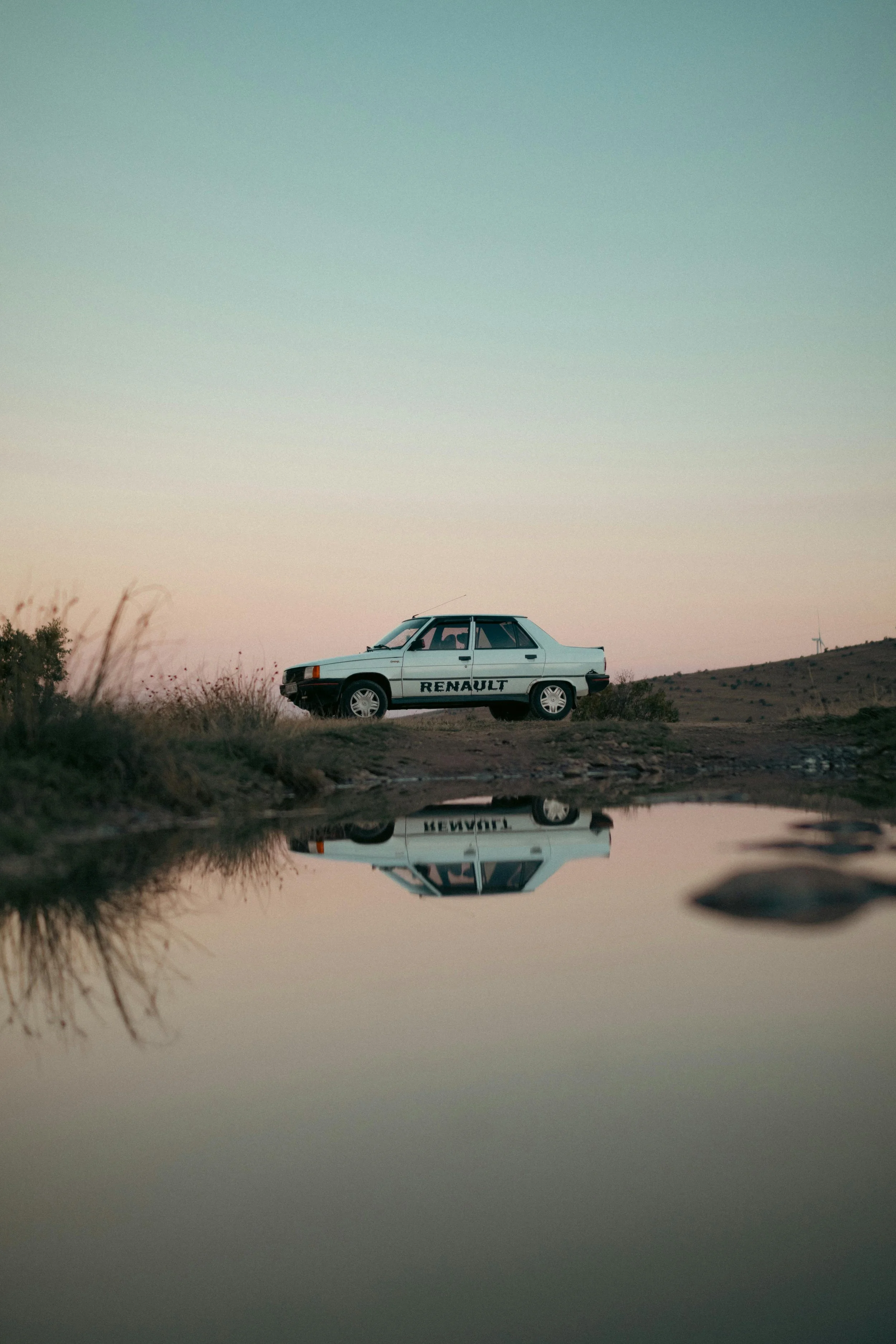 A vintage Renault car parked on a hill at sunset with its reflection visible in a puddle of water in the foreground.