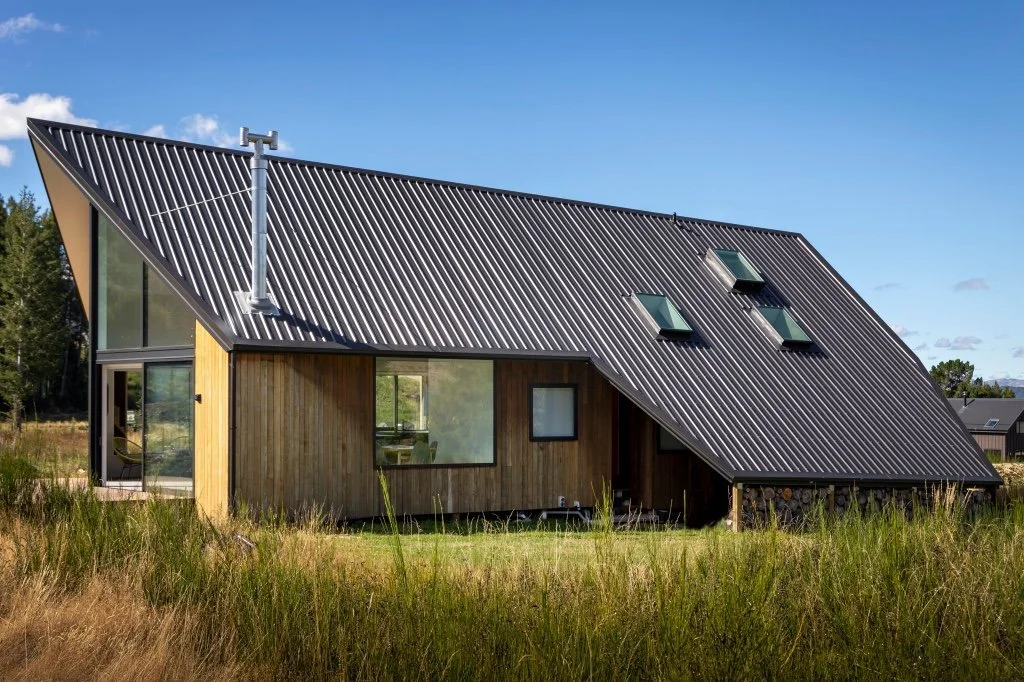 Modern house with slanted metal roof, wooden exterior, and large windows, set in a grassy landscape.