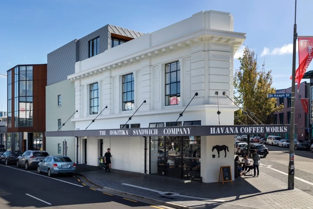Street view of The Hokitika Sandwich Company and Havana Coffee Works in a corner building with parked cars and people sitting at outdoor tables.