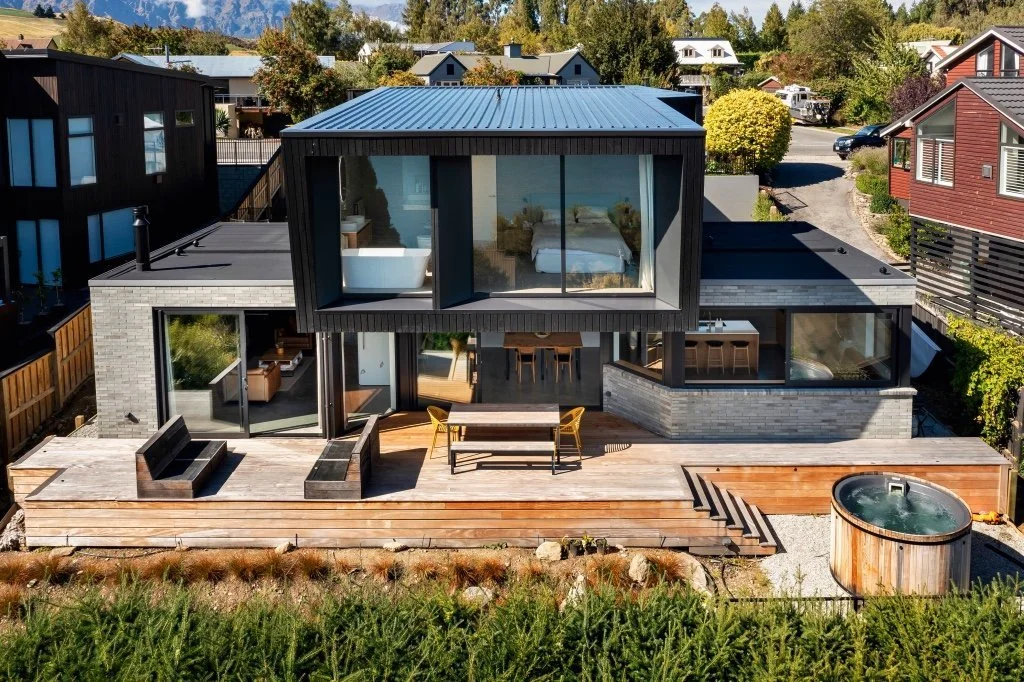 Modern two-story house with large windows, wooden deck, and circular hot tub in a residential area.