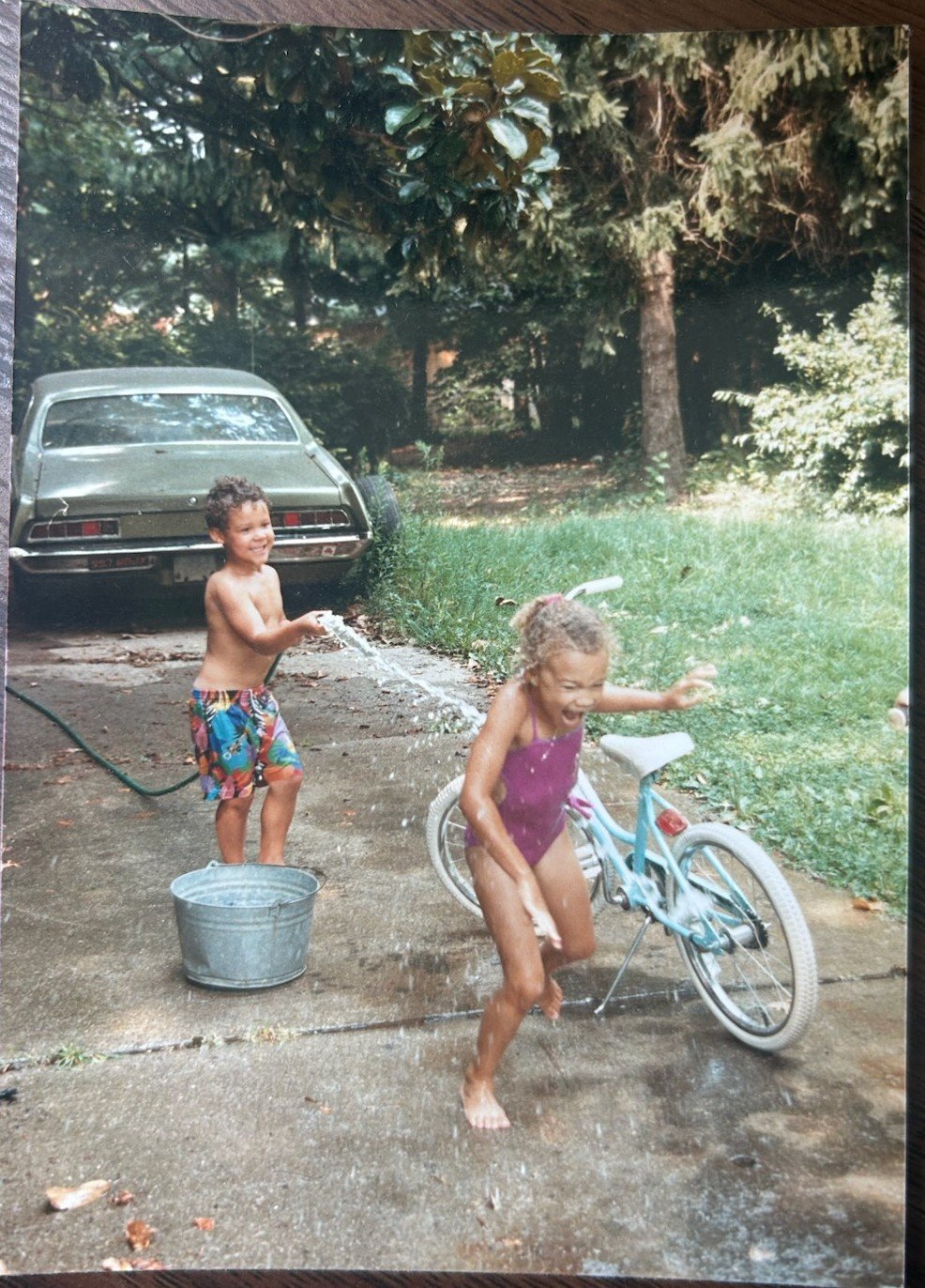A child outdoors beside a parked car in a wooded yard.