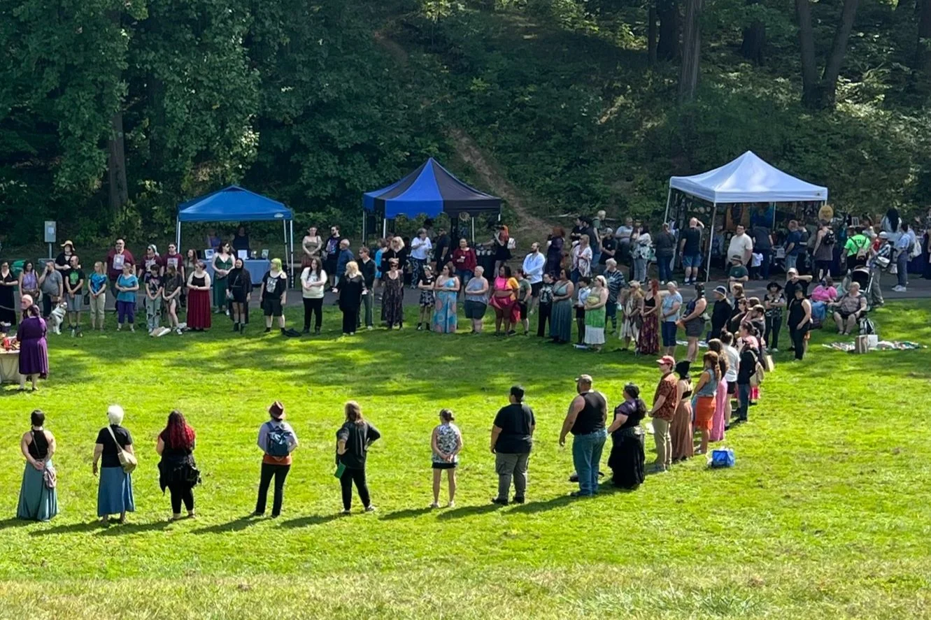 A large group of diverse people stand in a grassy area in a ritual circle, vendor tents in the background at 2024 CNY Pagan Pride Day