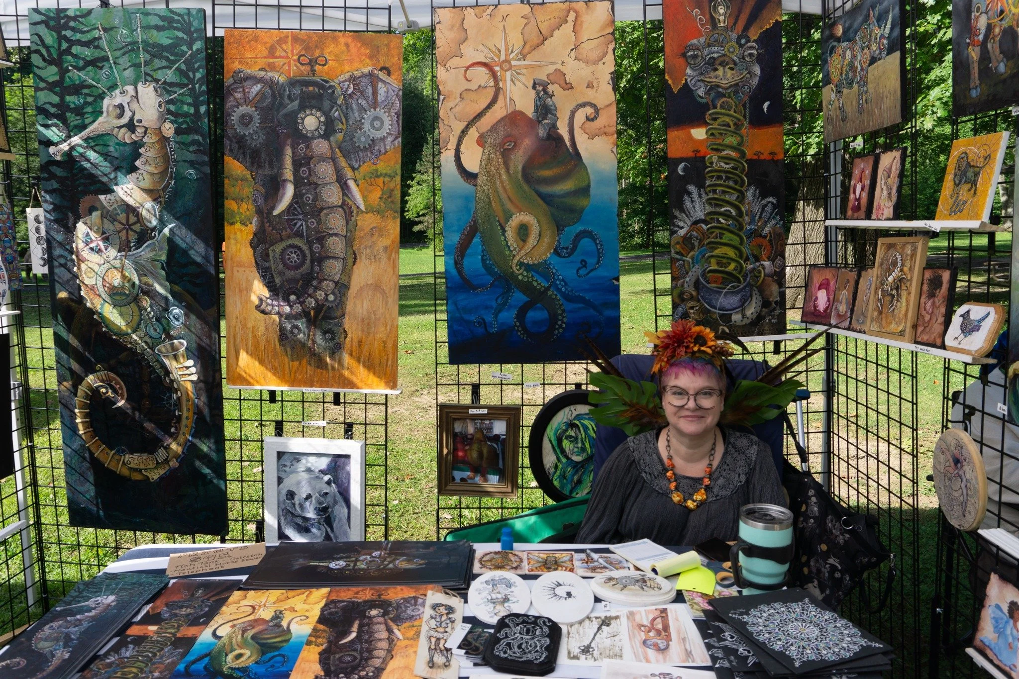 Steampunk and animal inspired paintings hang in an outdoor tent at 2023 CNY pagan pride day. The artist sits behind a table