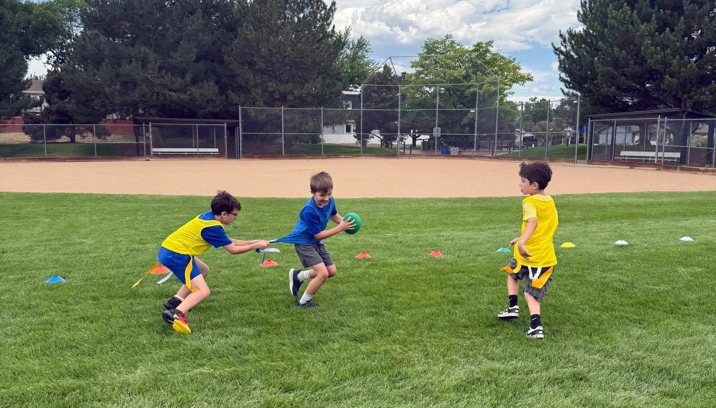 Three boys playing tug-of-war with a ball on a grassy field. Two are wearing yellow and blue sports uniforms, and one is wearing a blue shirt and gray shorts. There are small colored cones on the grass, and a baseball diamond with a fence in the back