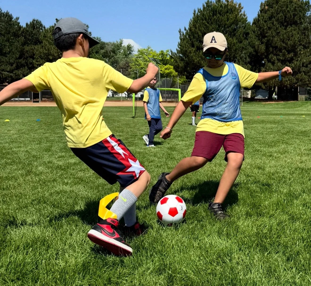 Two kids playing soccer on a grassy field with trees and a small goal in the background, one kid in a yellow shirt and red shorts, the other in a yellow shirt with a blue vest and maroon shorts, both wearing caps and sneakers, actively competing for