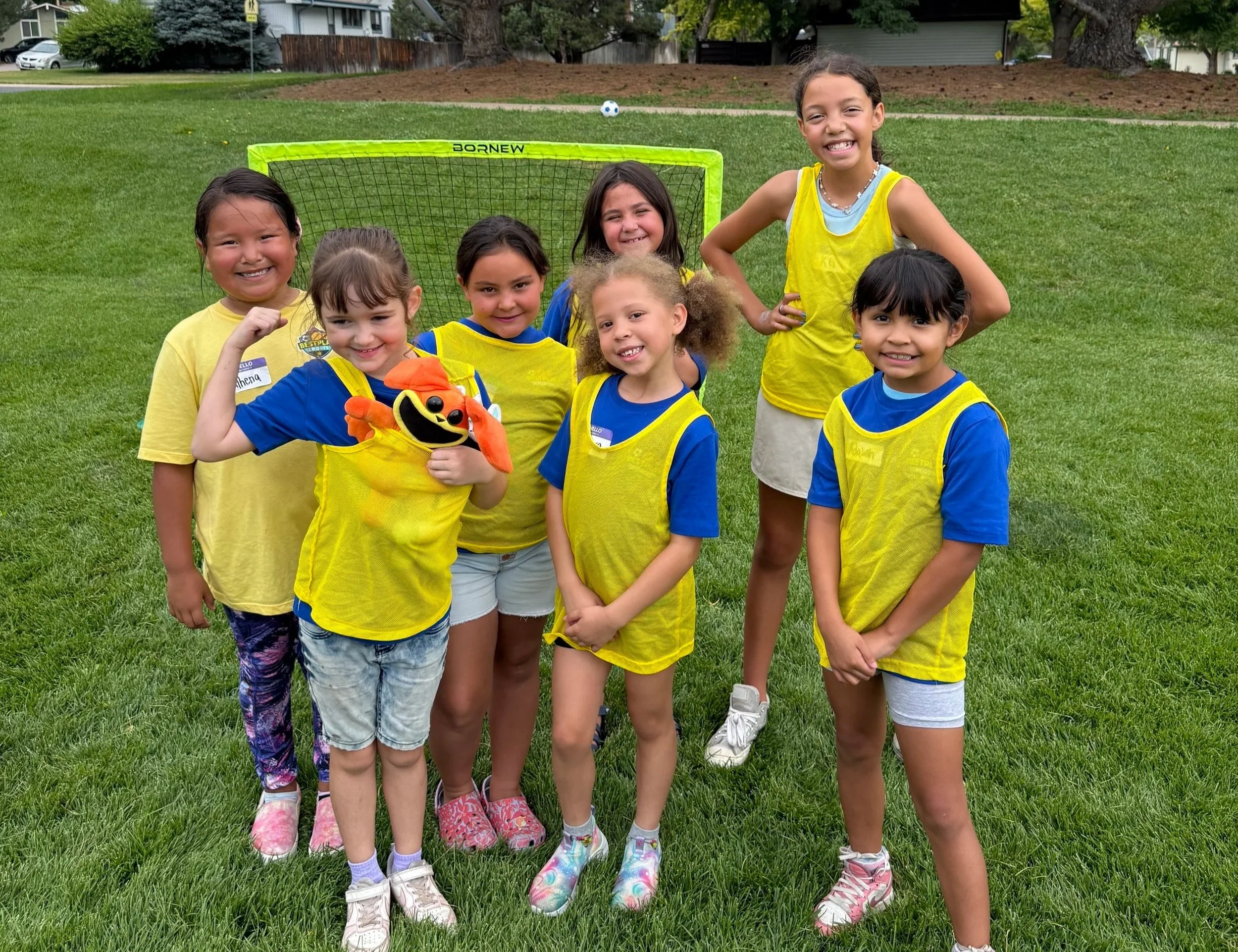 Group of seven young girls in yellow and blue sports jerseys standing on a grassy field with a small soccer goal and a soccer ball in the background. They are smiling, with one girl flexing her arm and another holding a plush toy puppet.