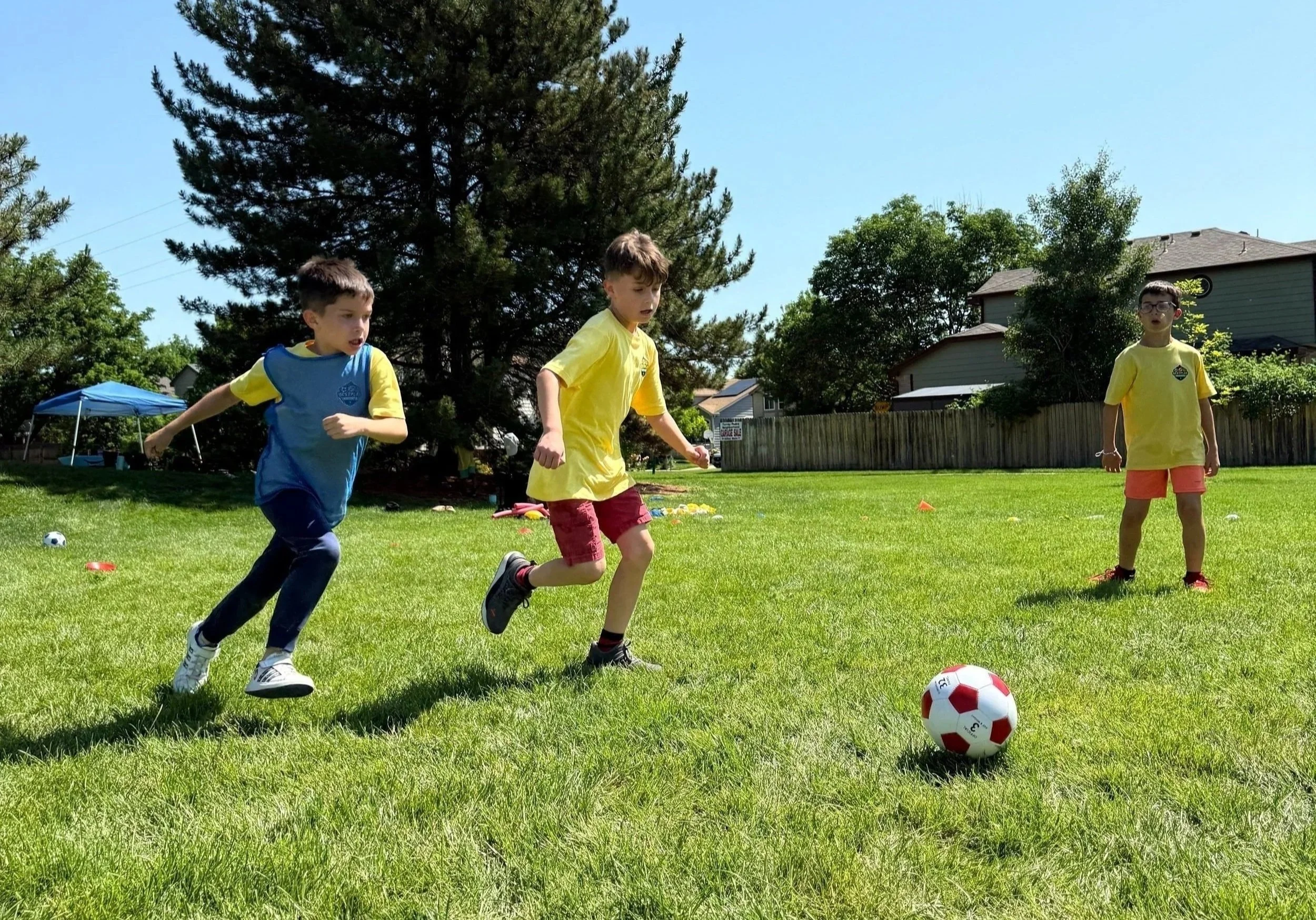 Three young boys playing soccer on a green lawn in a residential neighborhood on a sunny day. One boy is running, another about to kick the soccer ball, and the third standing nearby.