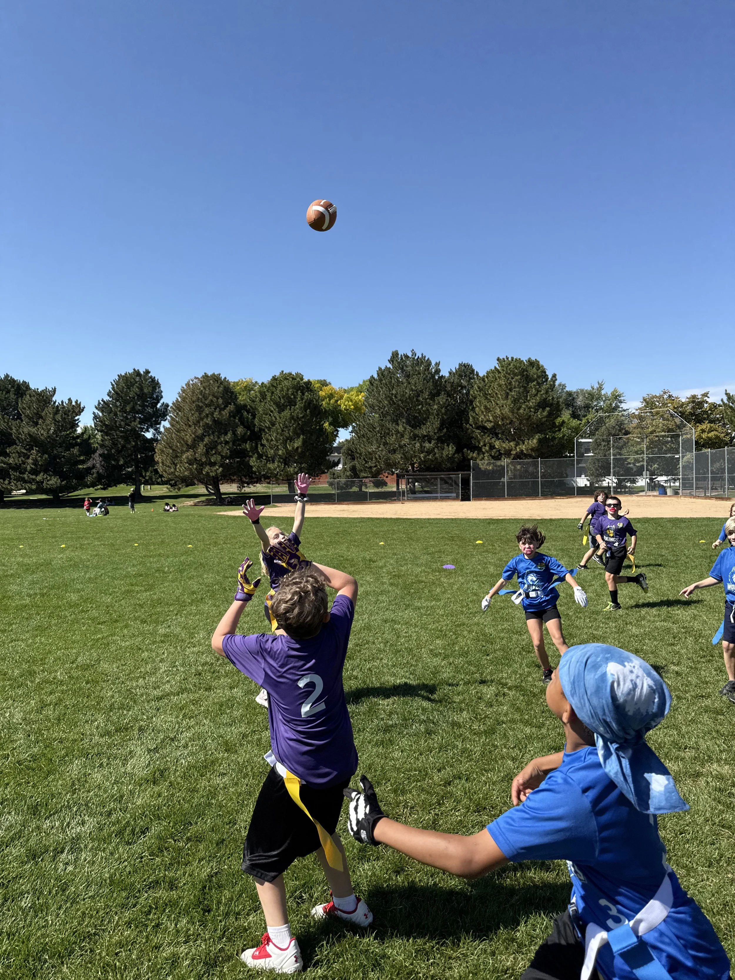 Children playing flag football on a grassy field during a sunny day, with some kids running and others reaching for the football in mid-air. Bestplay Sports Colorado.