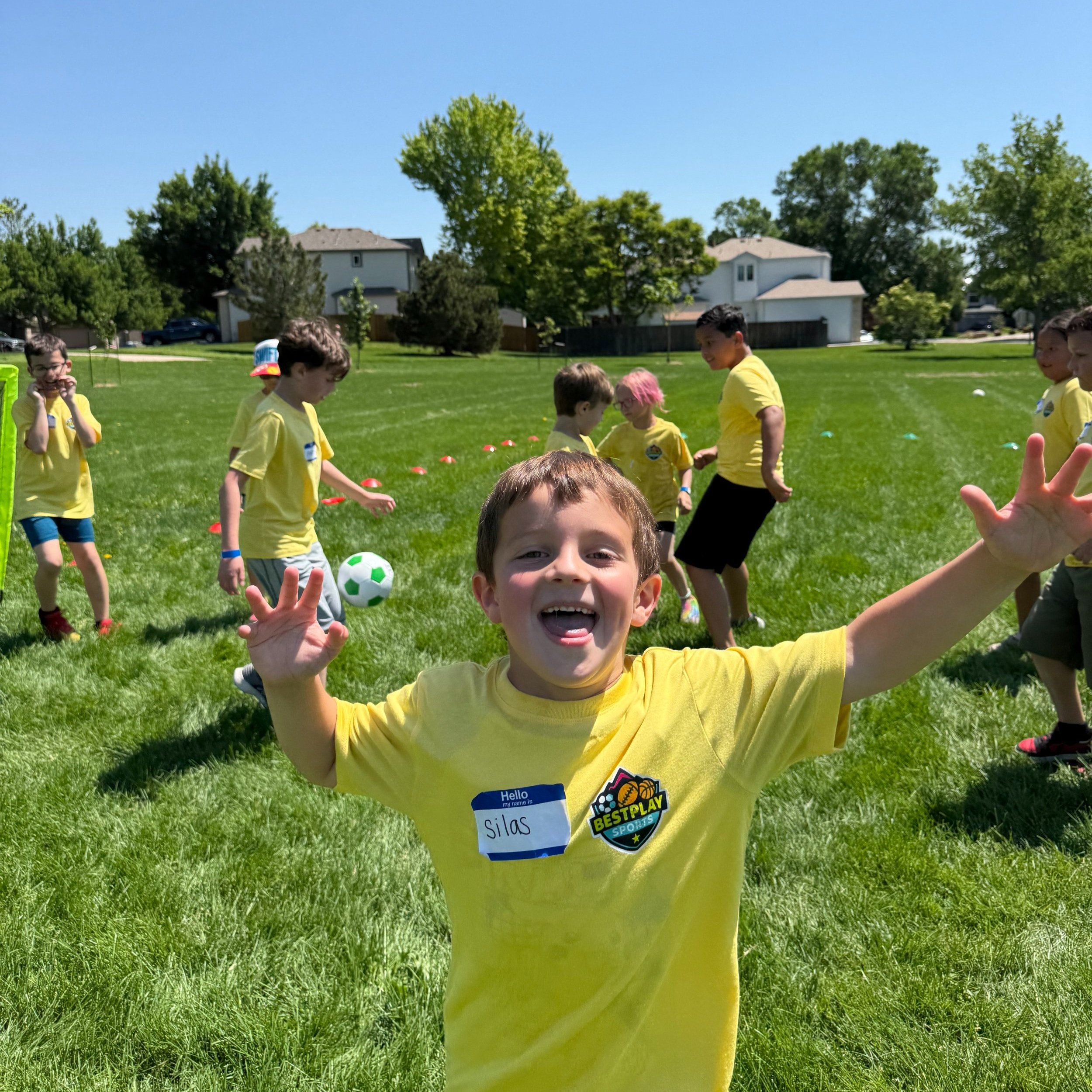 A young boy in a yellow shirt smiling and making a peace sign with his hands in a park during a soccer game with kids in yellow shirts playing in the background.