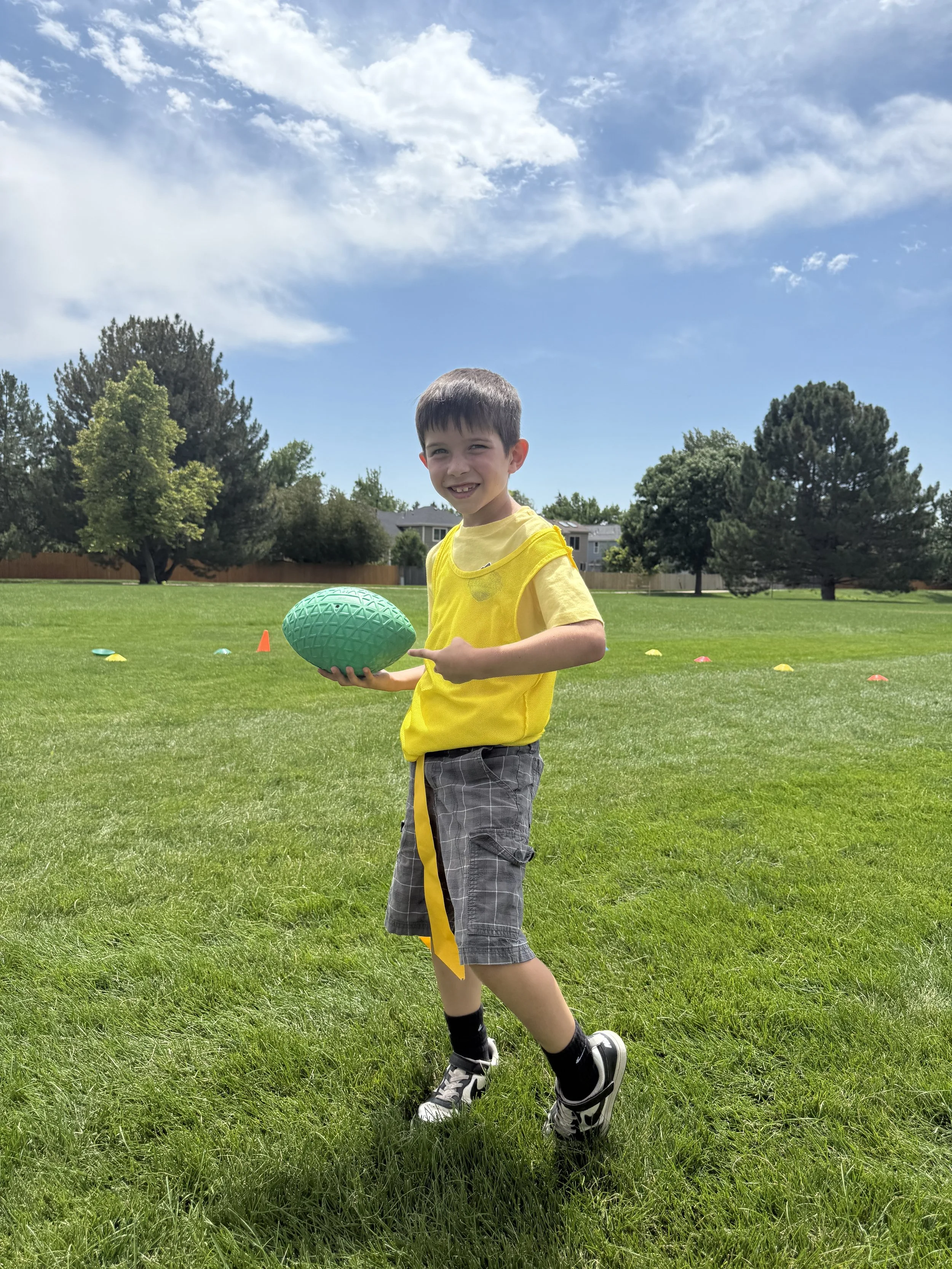 A young boy wearing a yellow shirt and gray plaid shorts holding a green football on a grassy field, with trees and a blue sky with clouds in the background.