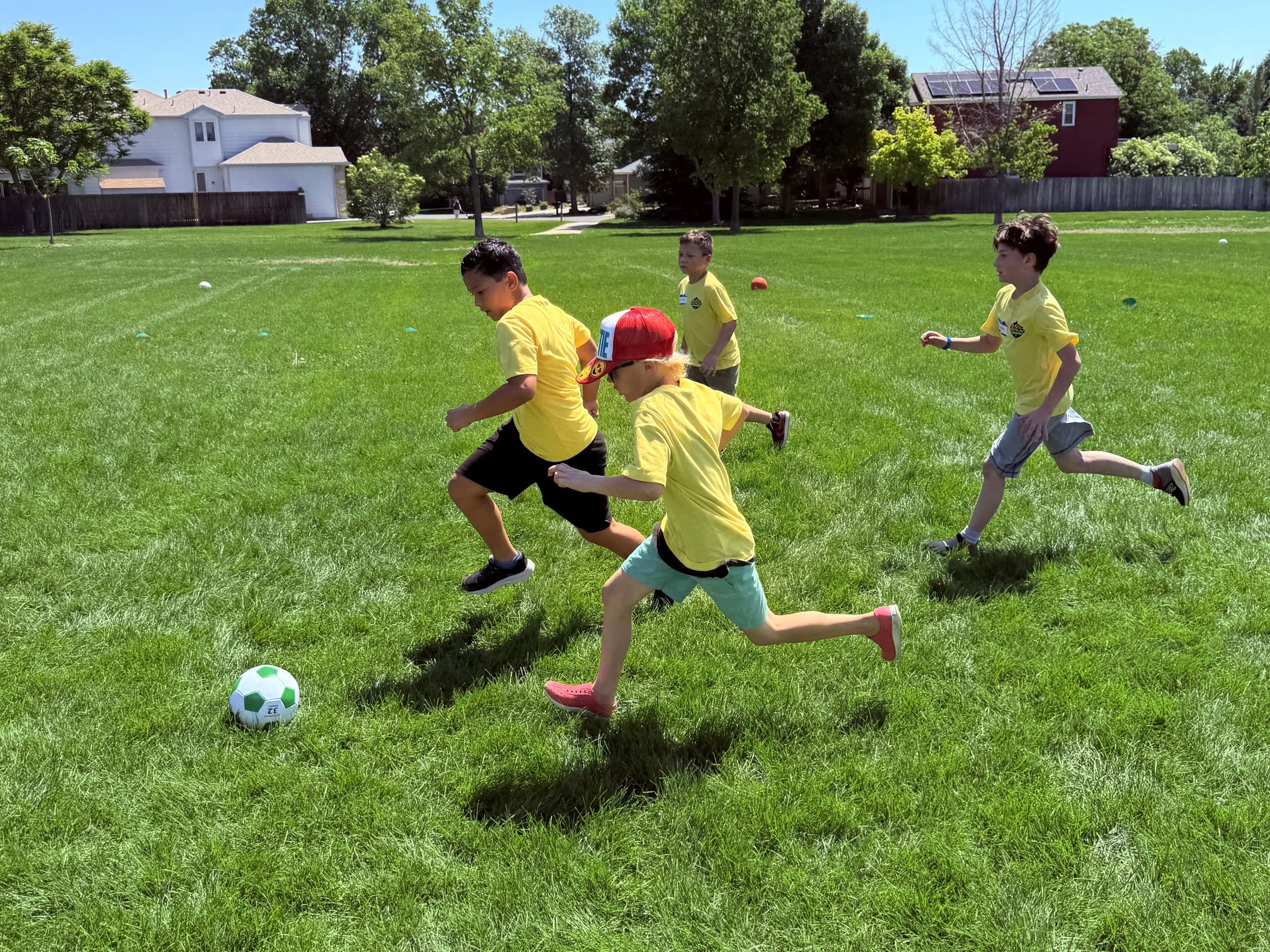 Four children wearing yellow shirts playing soccer on a grassy field on a sunny day.