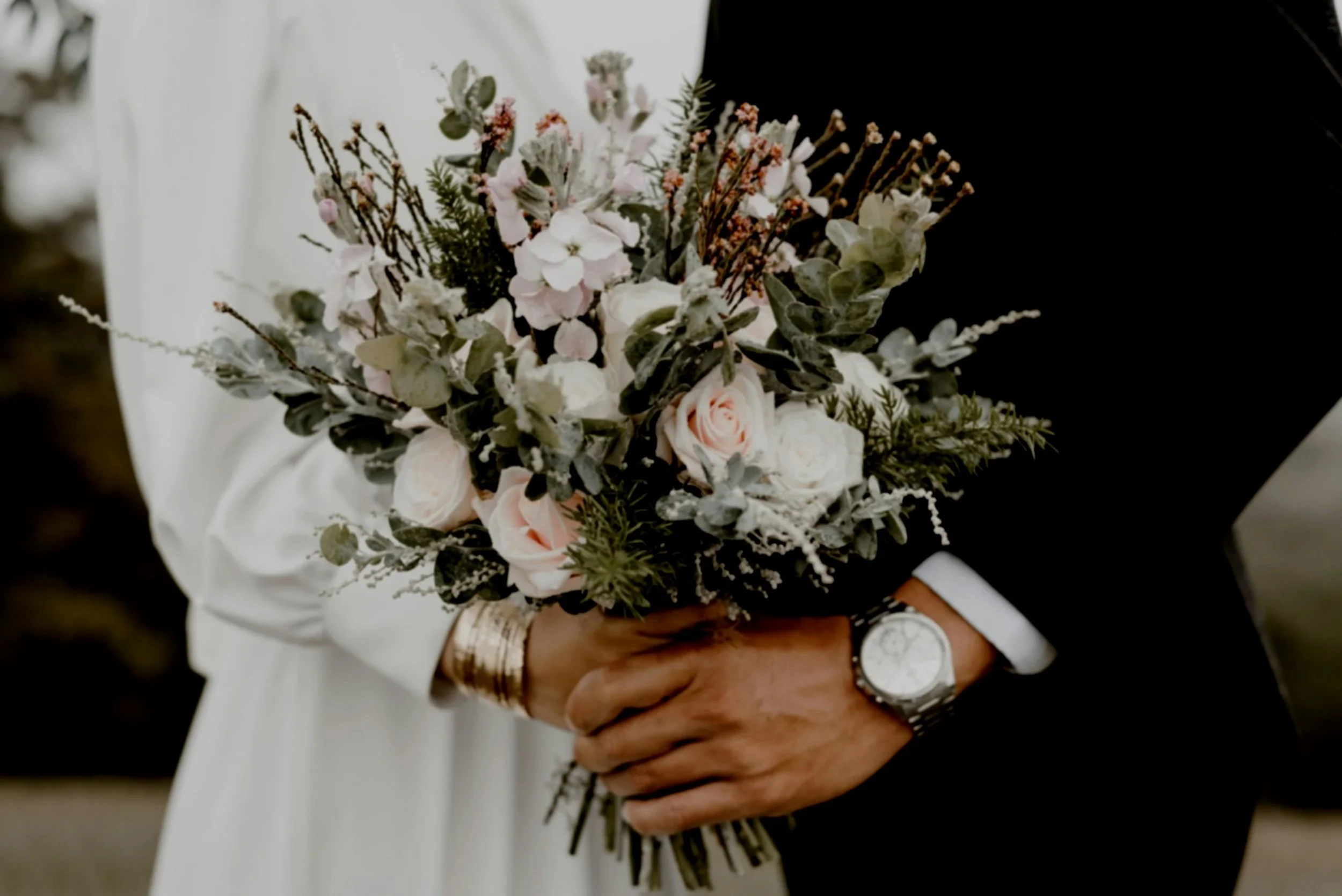 A couple holding a wedding bouquet with white and pink roses, greenery, and delicate flowers, with the groom wearing a black suit and a silver watch.