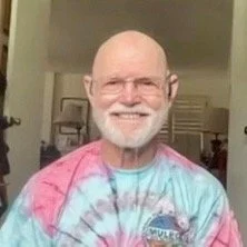 An older man with a white beard and glasses, smiling, wearing a tie-dye shirt.