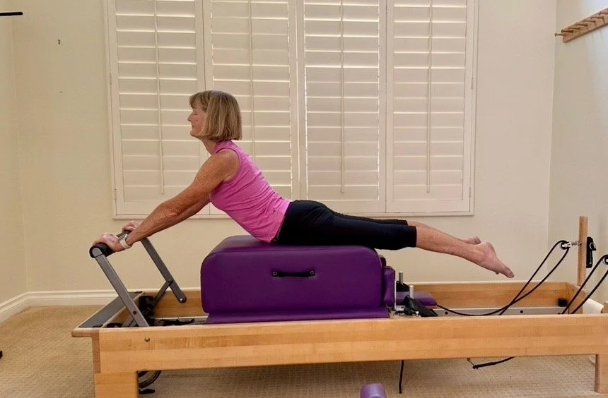 A woman performing an exercise on a Pilates reformer machine, lying on her stomach with her arms extended forward and legs lifted straight back.