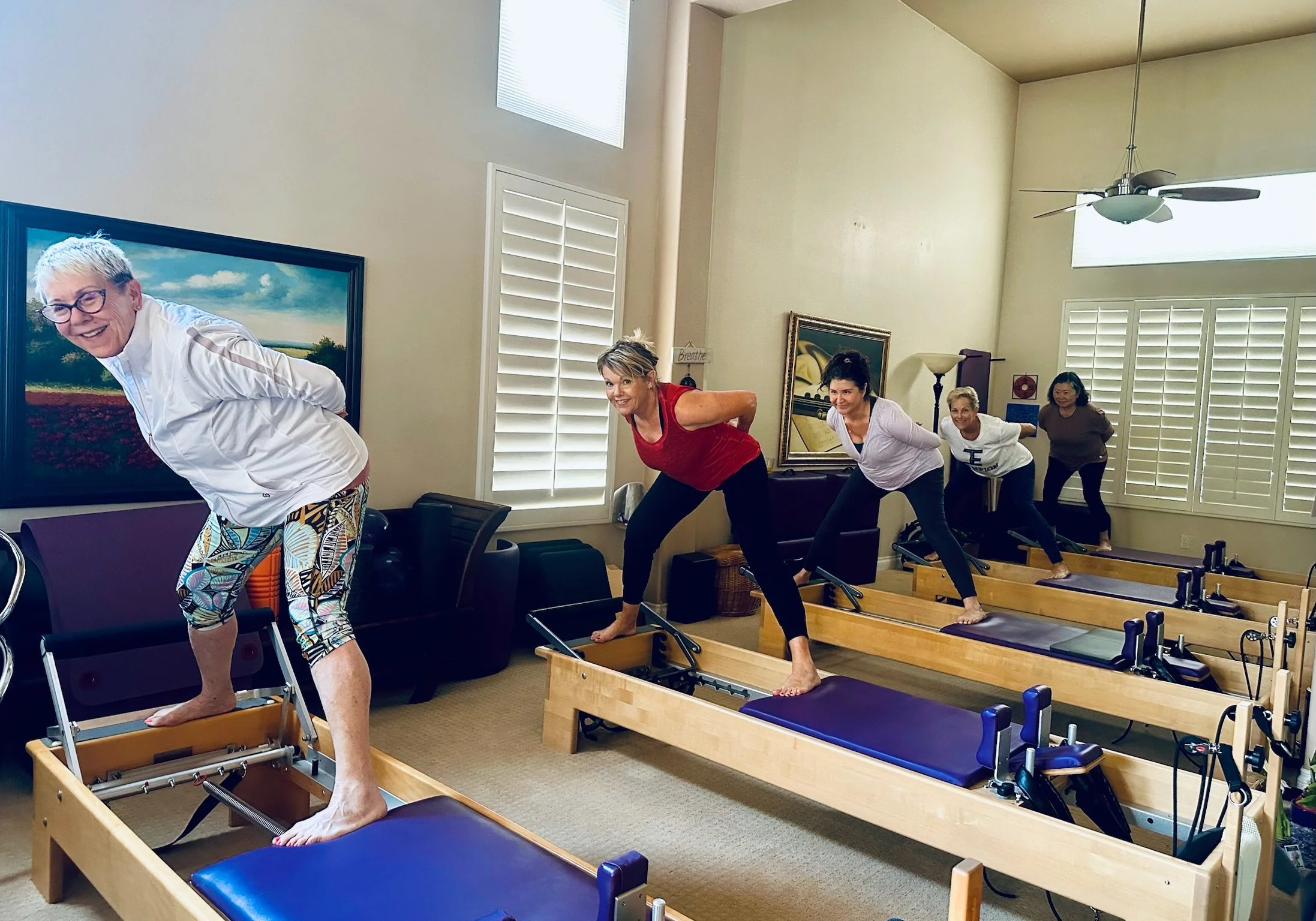 Group of women participating in a Pilates class on reformer machines in a bright living room with large windows and plantation shutters.