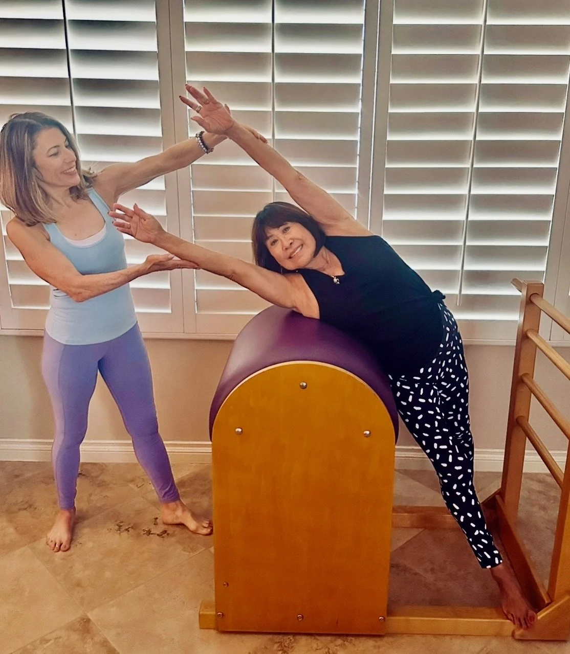 Two women practicing yoga indoors, one standing and holding the other's hand, who is leaning on a wooden barrel with her arm extended and a smile. Behind them are white shutters.