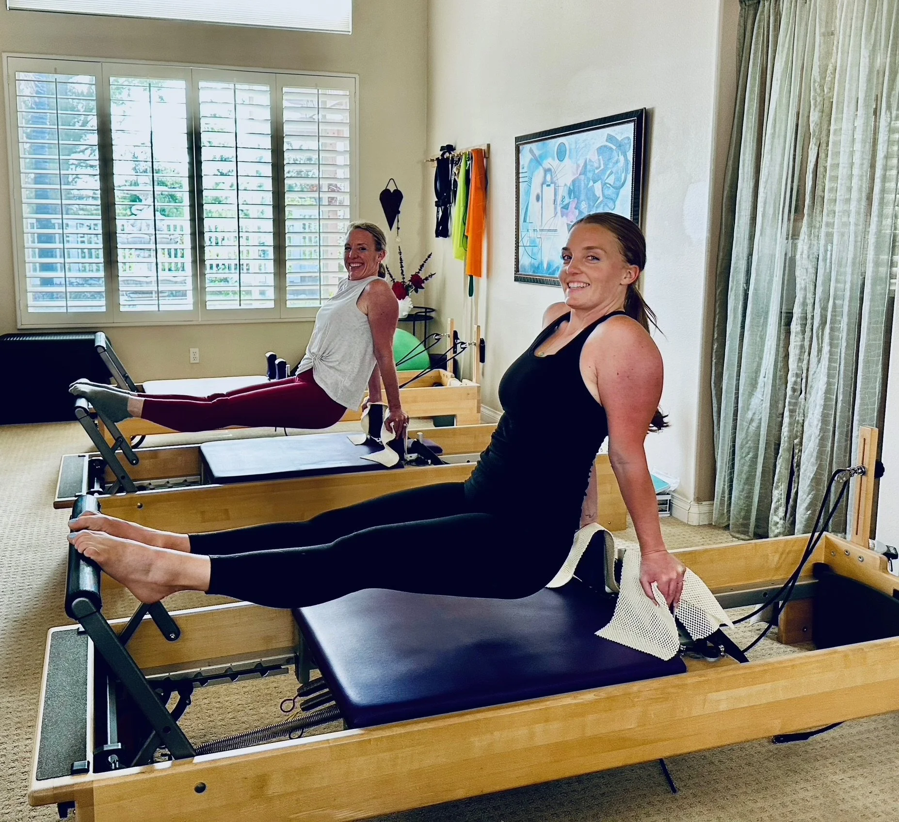 Two women using Pilates reformer machines in a well-lit room with large diagonal windows, smiles, and relaxed postures.