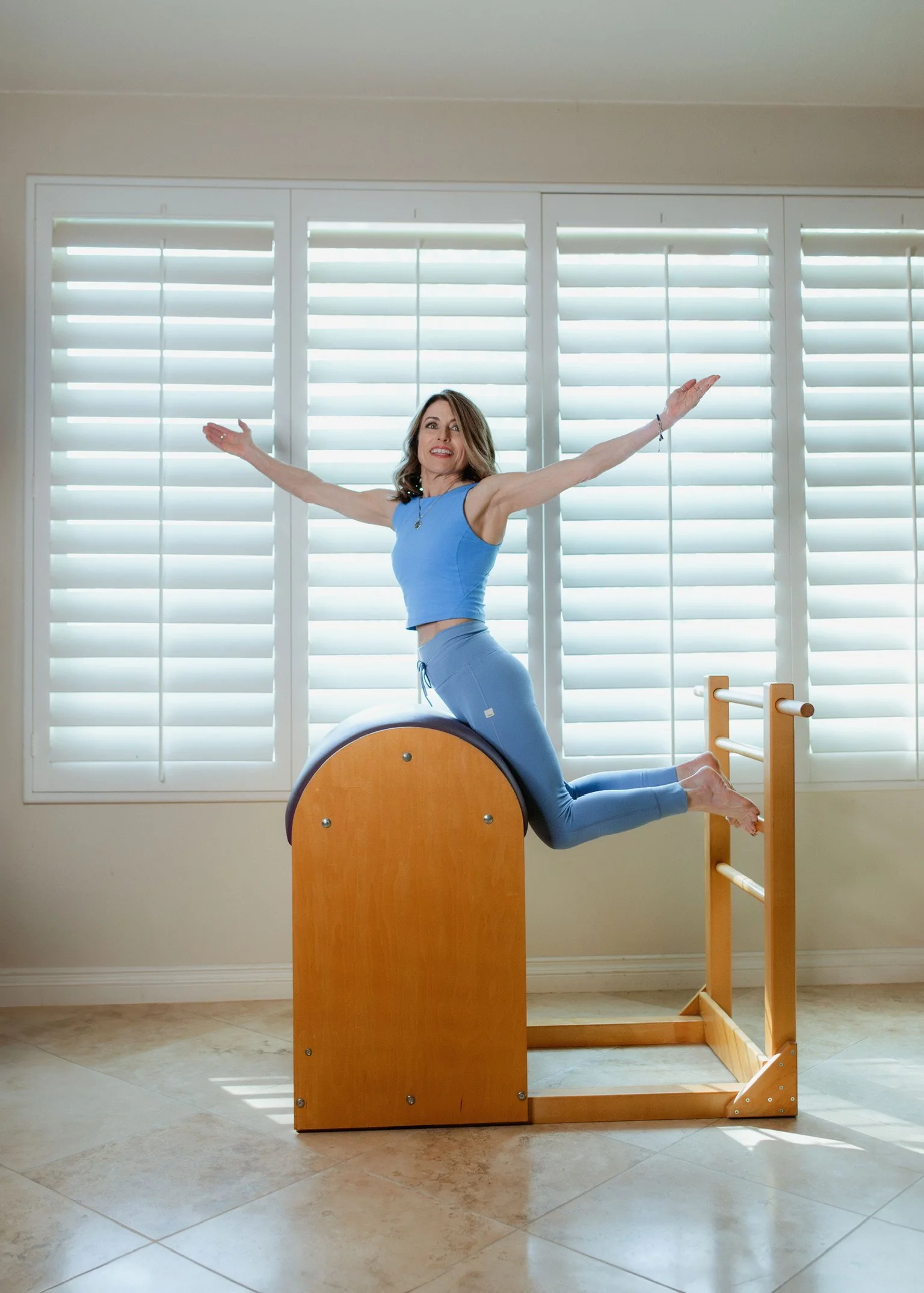 Woman performing a Pilates exercise on a wooden Pilates barrel with a large window and white blinds in the background.