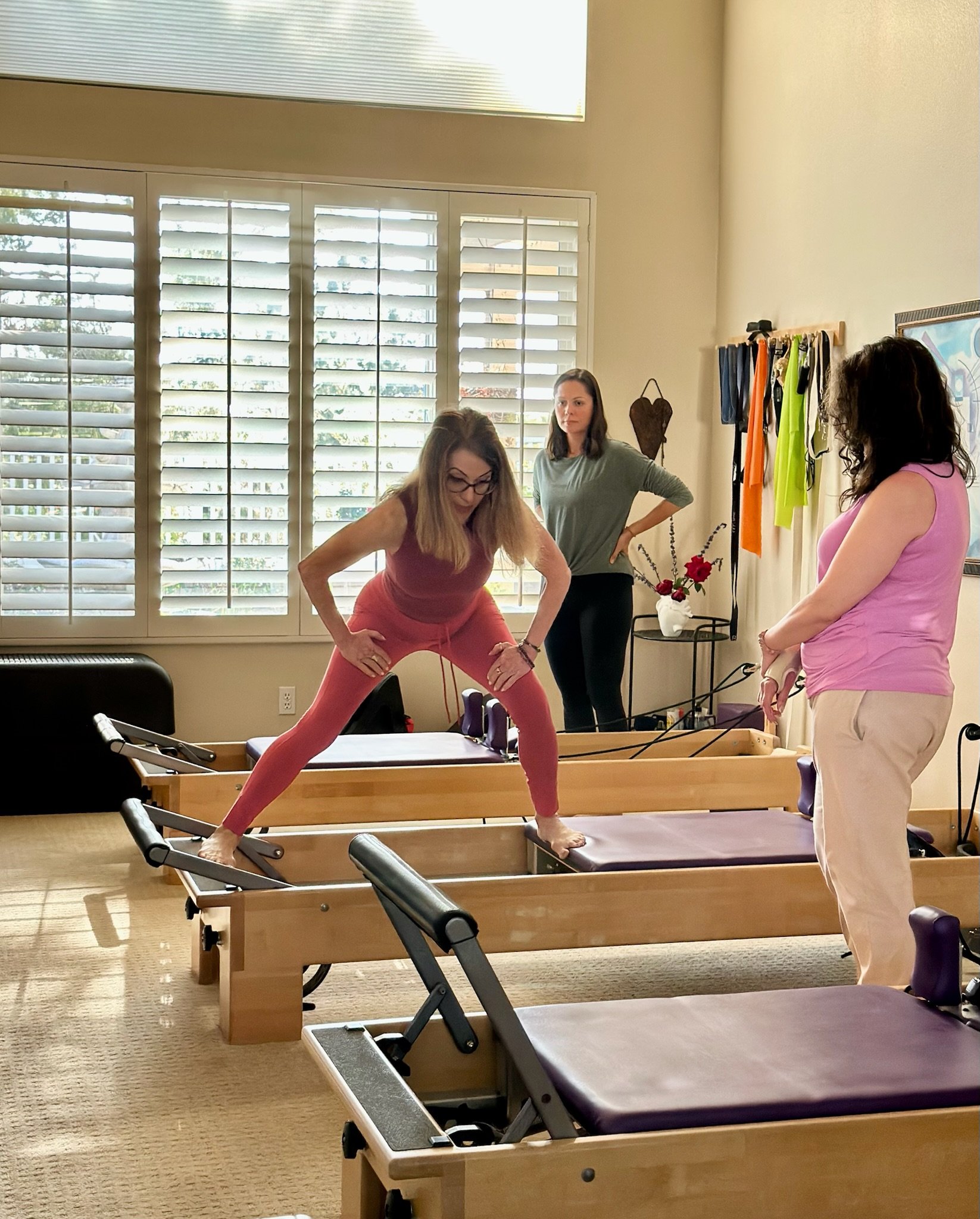 A woman in pink workout clothes is balancing on a Pilates reformer machine while two women observe her. The room has large windows with shutters, exercise equipment, and colorful fitness accessories on racks.