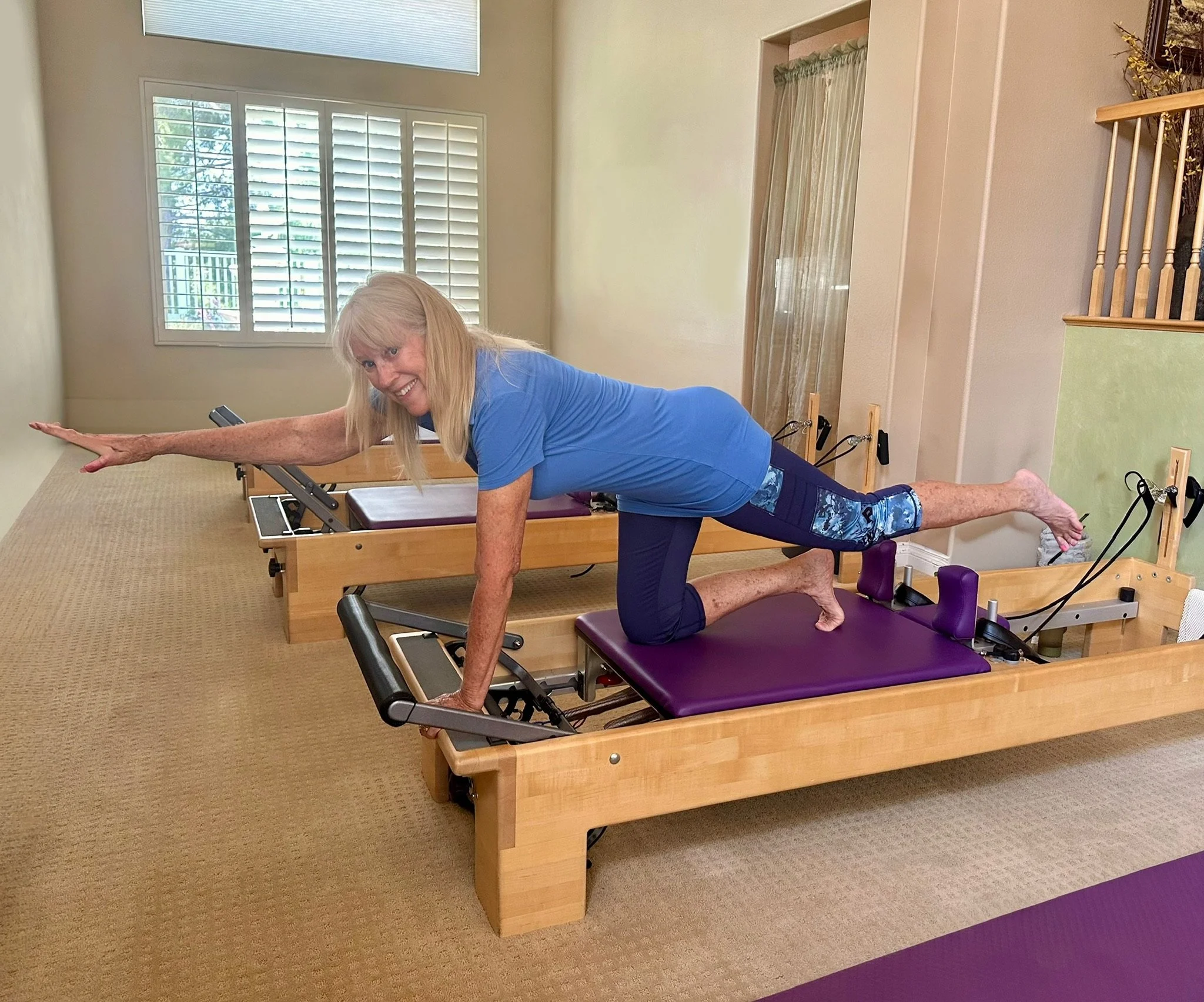 An elderly woman doing Pilates on reformer machines in a bright room with a window and beige walls.
