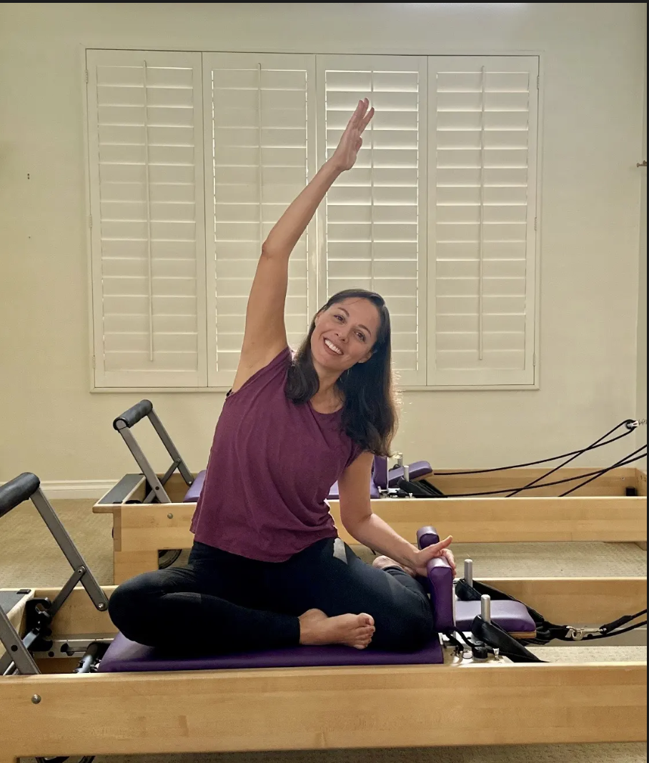 A woman practicing Pilates on a reformer machine, sitting cross-legged on a purple cushion, smiling, with her right arm raised and her left hand holding a handle, in a room with closed white shutters.