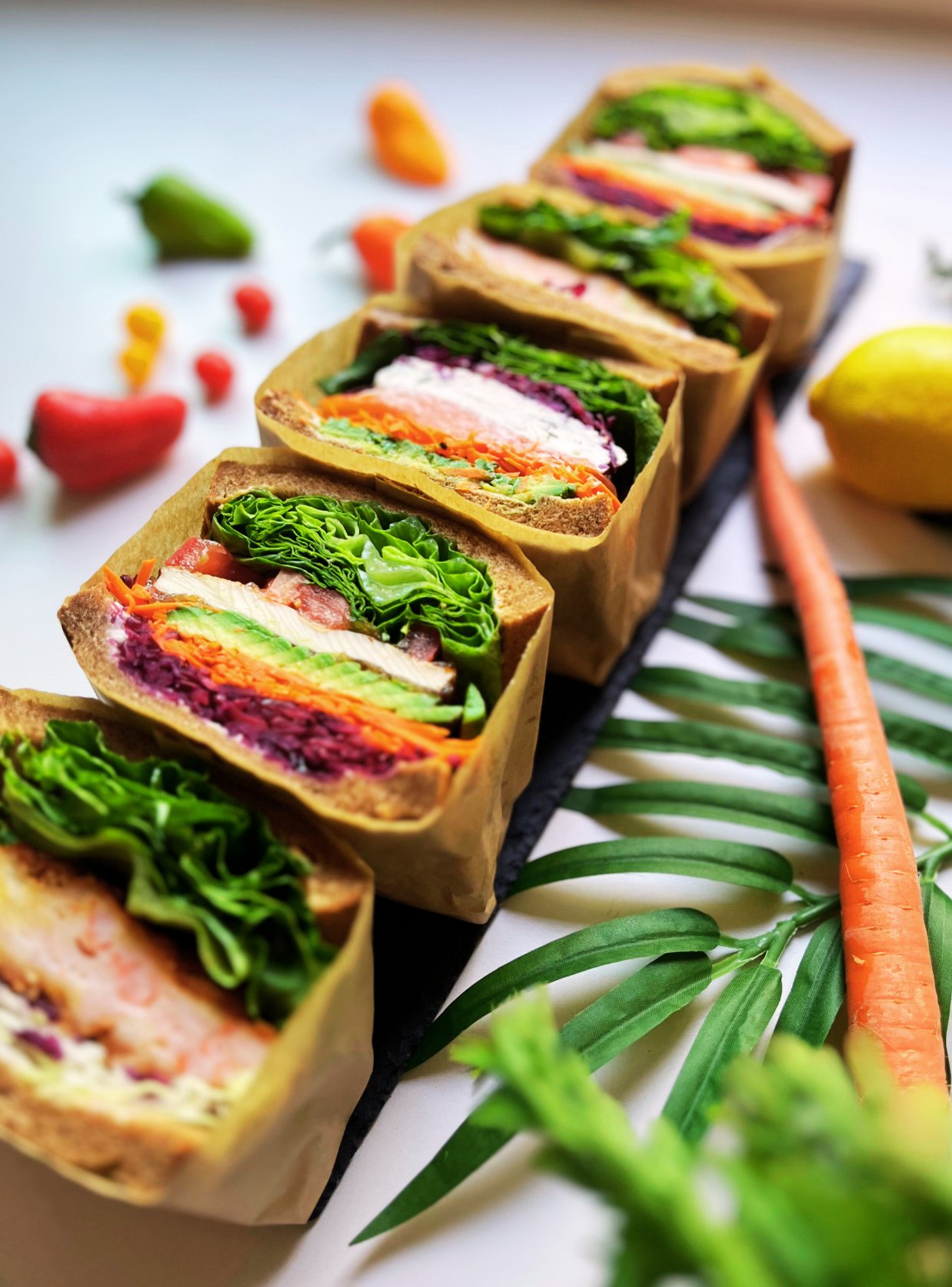 Line of colorful vegetable and hummus wraps on a black tray, with a carrot, lemon, and green leafy plant nearby, on a white background.