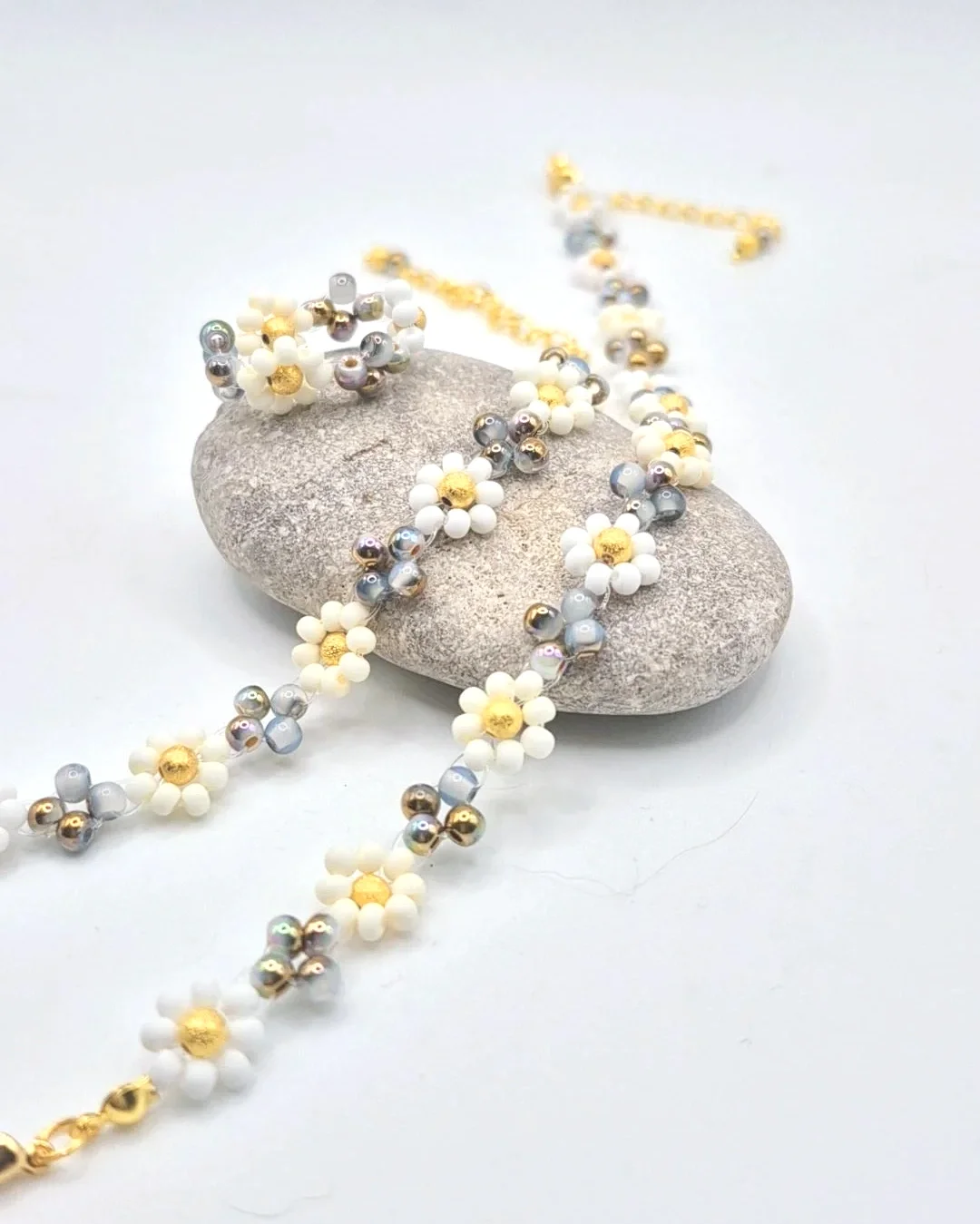 a set of handed-beaded jewlery imitating daisy flowers, including a bracelet, anklet, and ring, laid out on a river rock