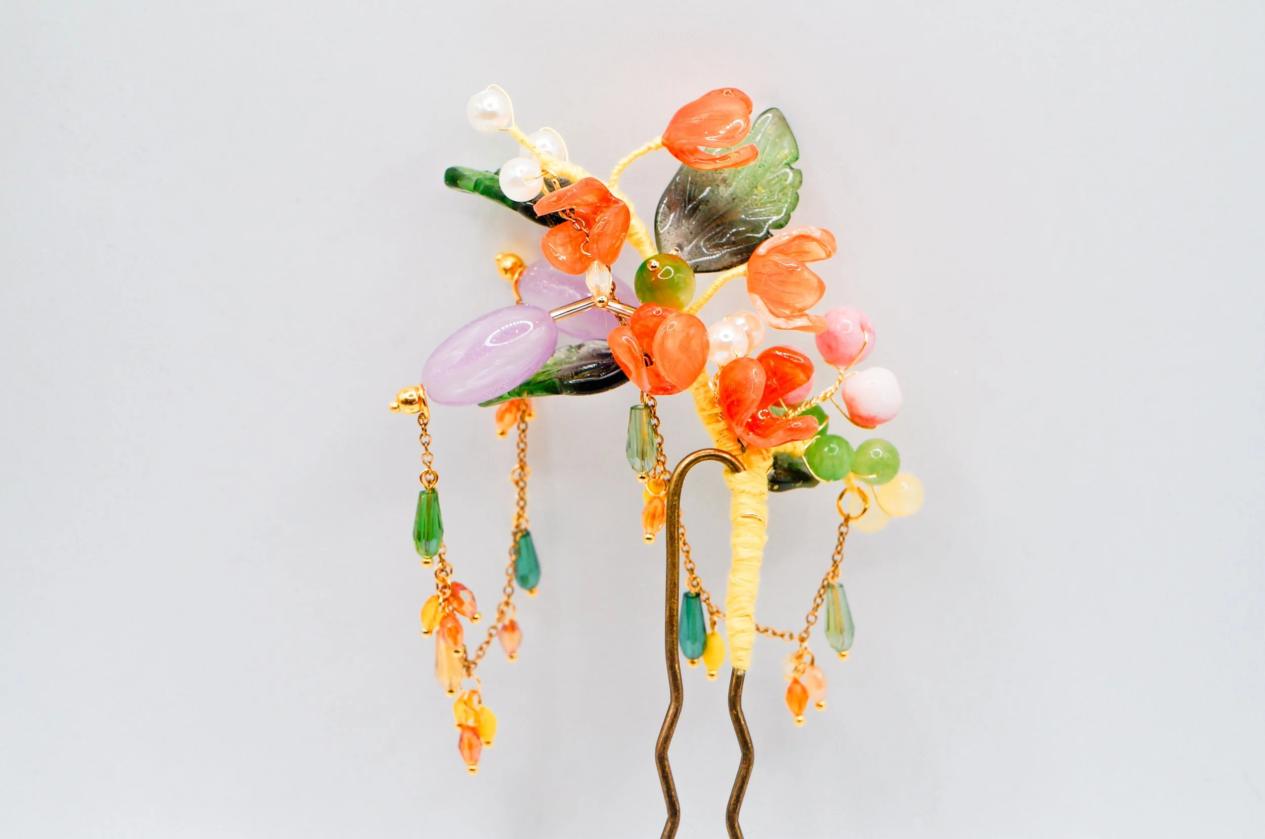Close up of the front of a hand crafted bronze U-shaped hairstick featuring orange blossom buds, gold speckled glass leaves, and dancing chains