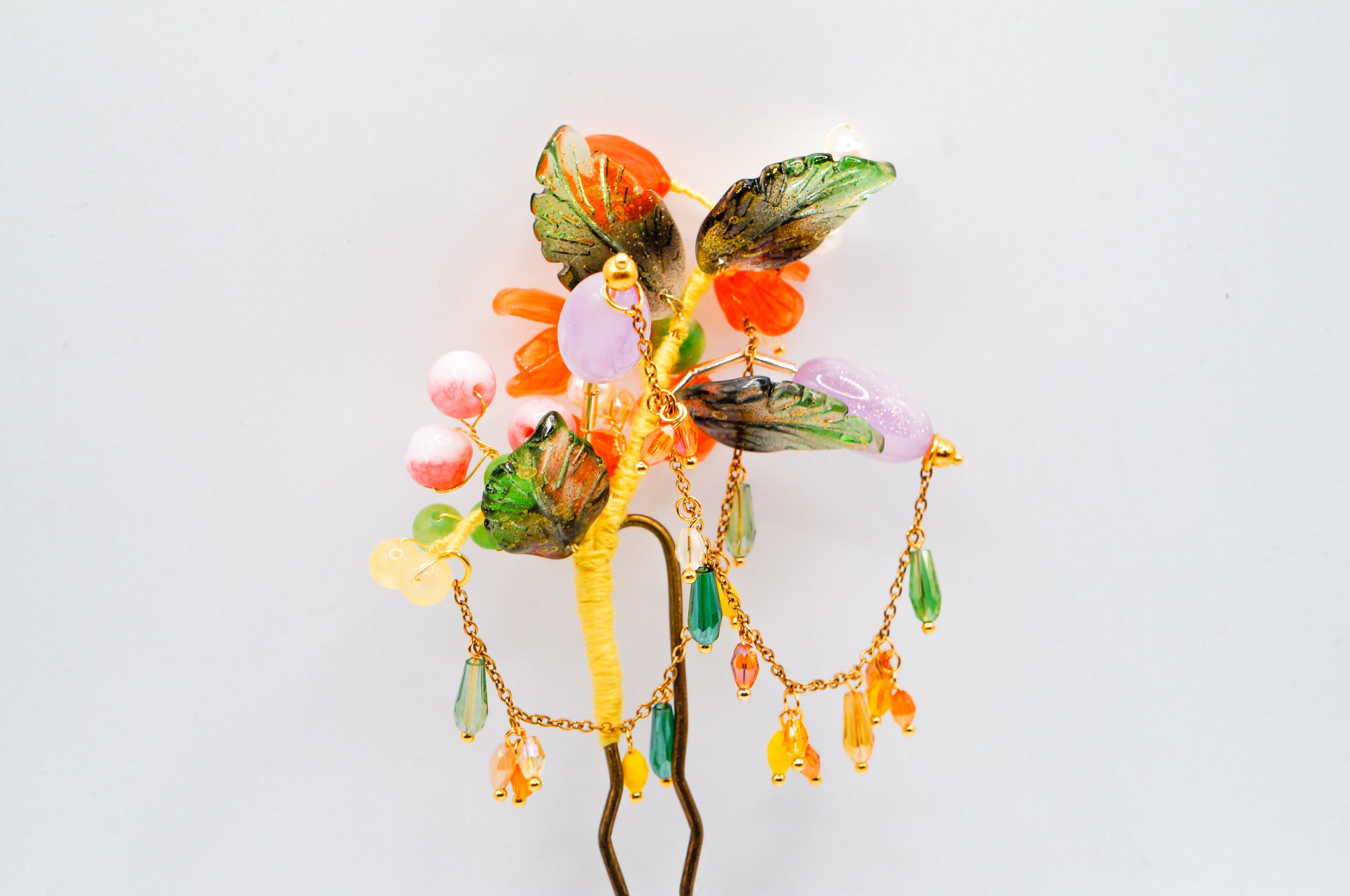 Close up of the backside of a hand crafted bronze U-shaped hairstick featuring orange blossom buds, gold speckled glass leaves, and dancing chains