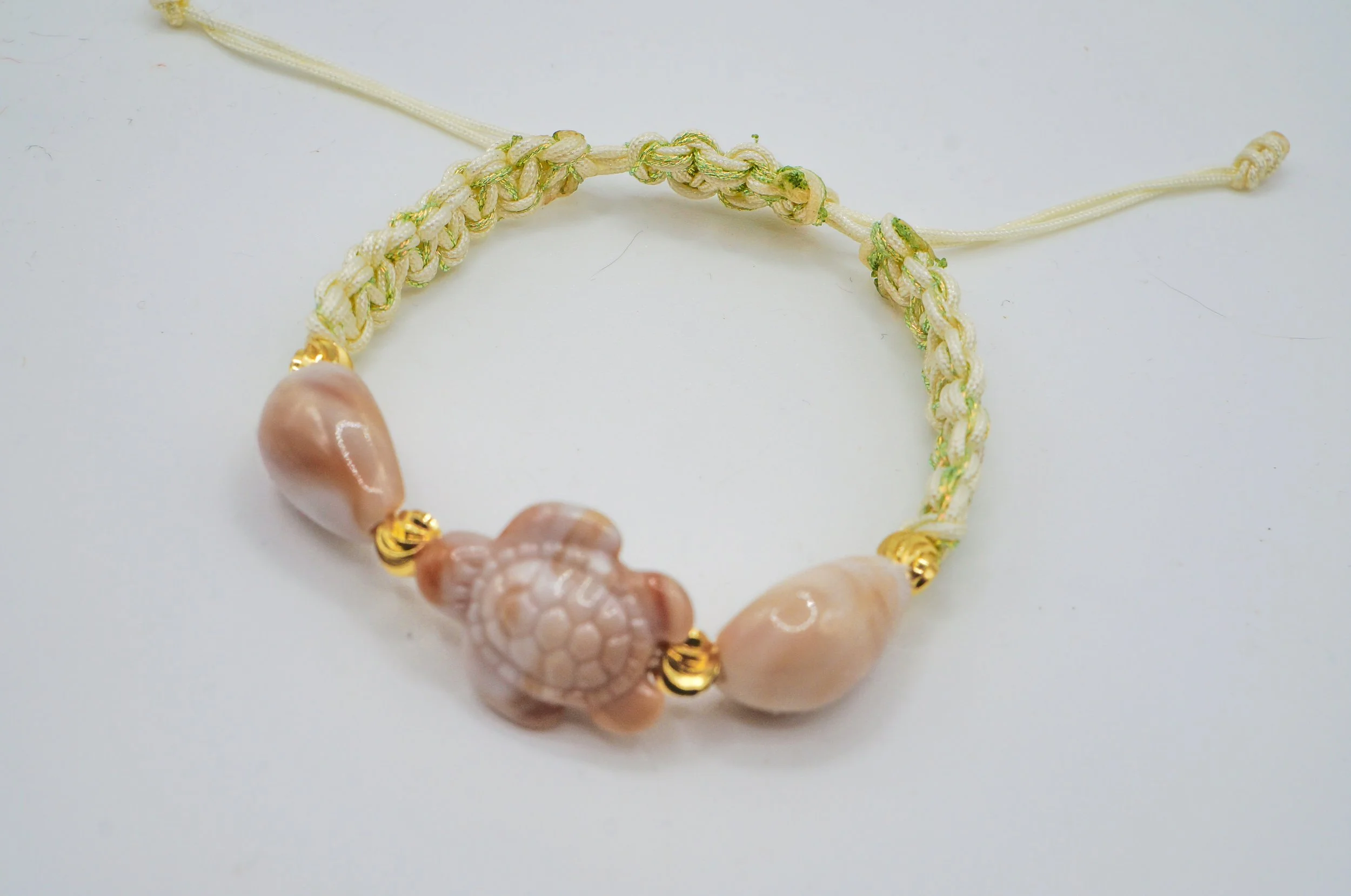 a white/metallic green double woven macrame bracelet with a turtle and two seashells, with gold spacer beads in between.