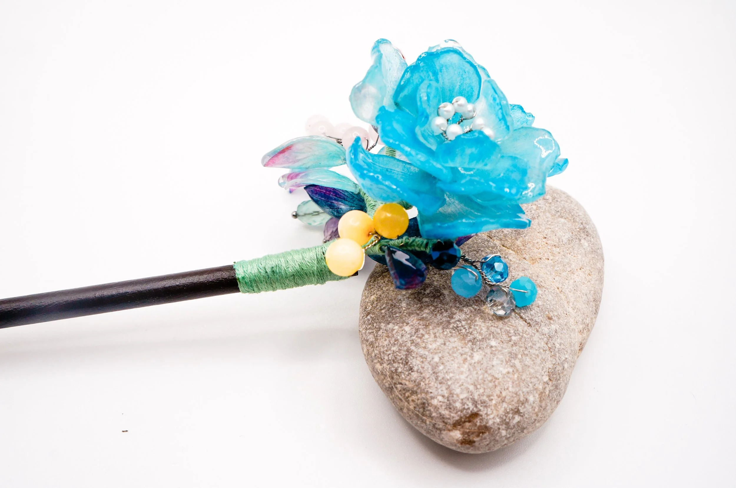 Close up of a handmade wooden chinese hair stick with large blue, multi-layered flower and various stone and glass beads with green silkthread wraps