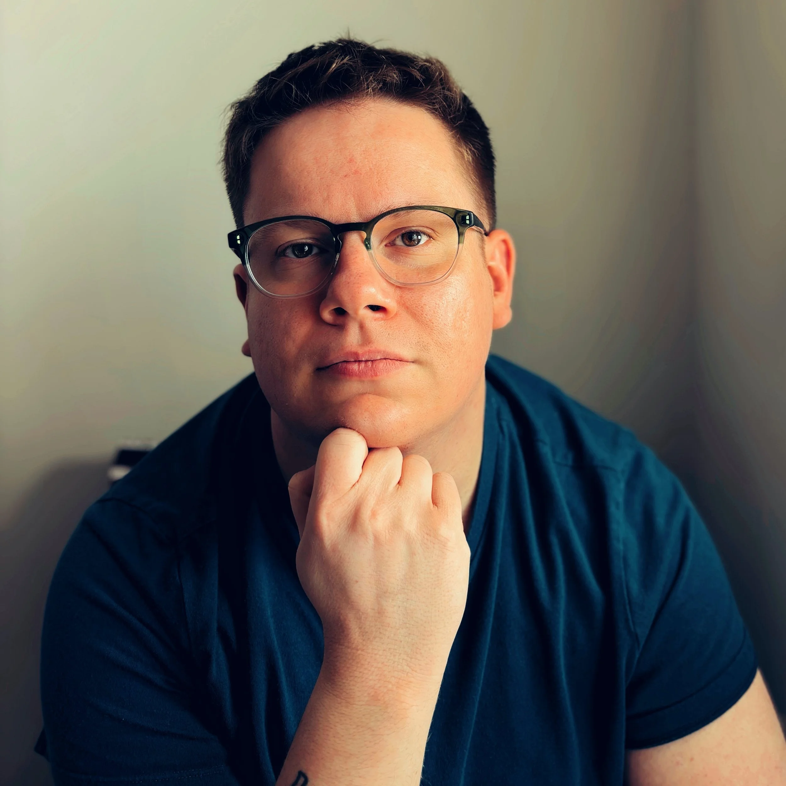 Portrait of a man with short dark hair wearing glasses and a blue shirt, resting his chin on his hand and looking serious.