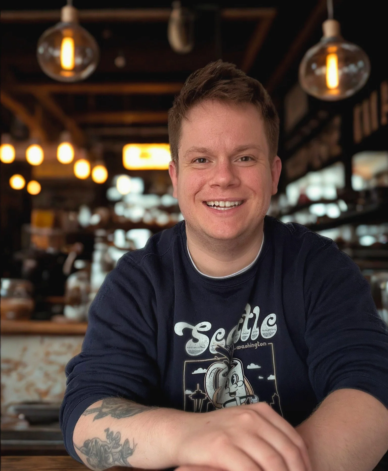 A smiling man with tattoos on his arm, wearing a Seattle-themed shirt, sitting in a warmly lit restaurant or cafe with hanging lights and blurred background.