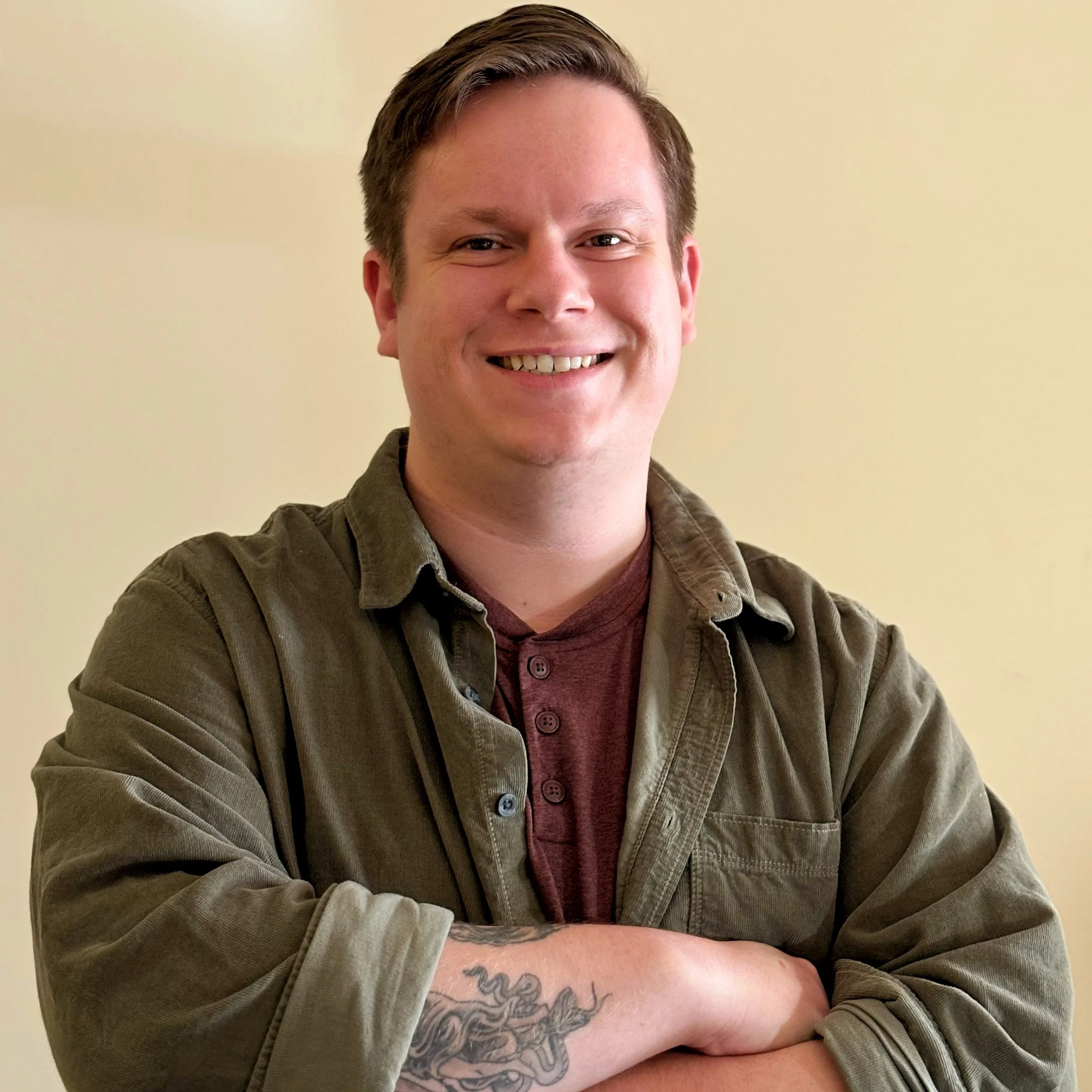 A smiling man with short brown hair wearing a green jacket over a maroon shirt, standing with arms crossed against a plain light-colored background.