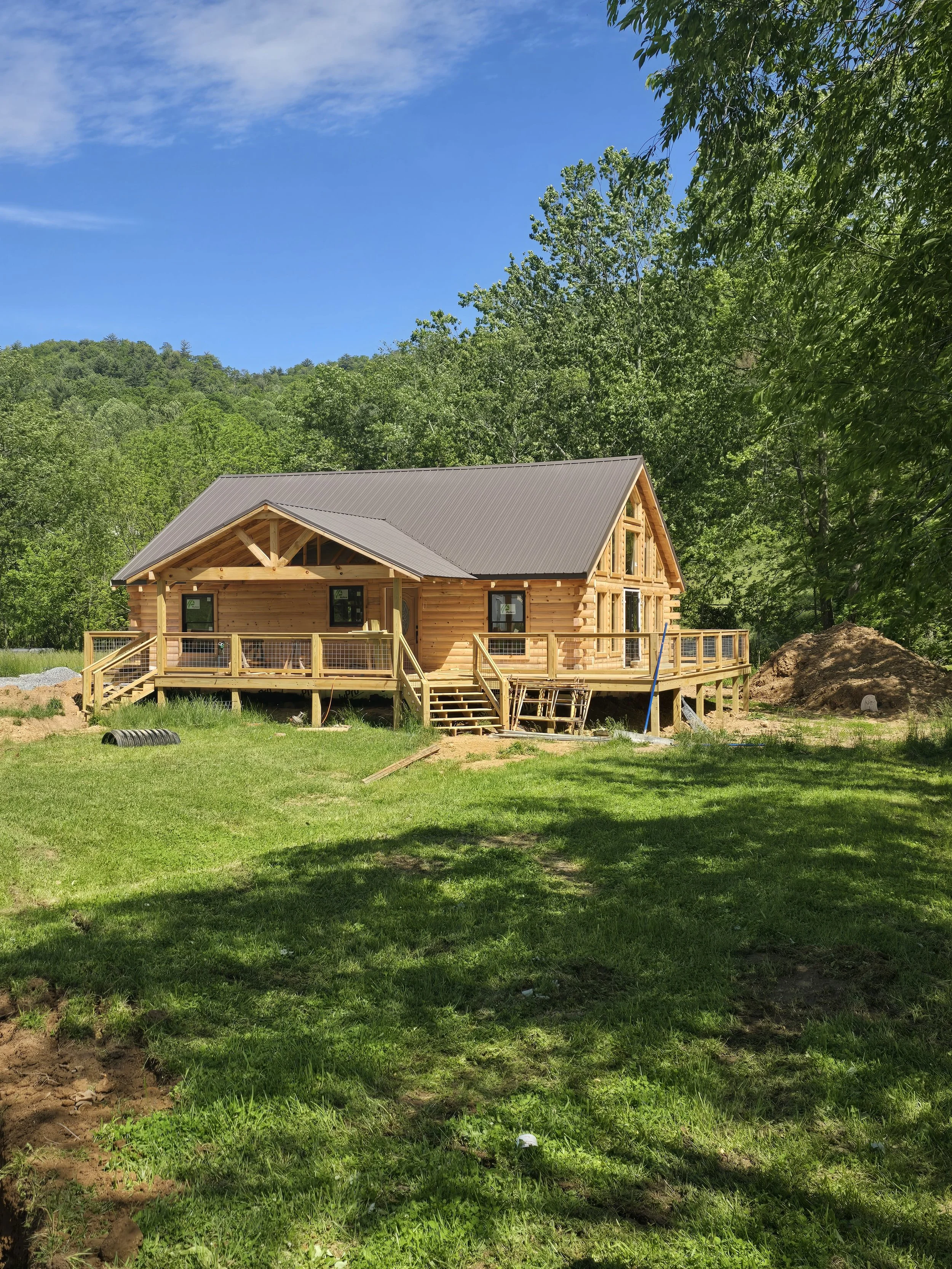 Newly built wooden house with a porch and stairs, surrounded by green grass and trees, under a blue sky.