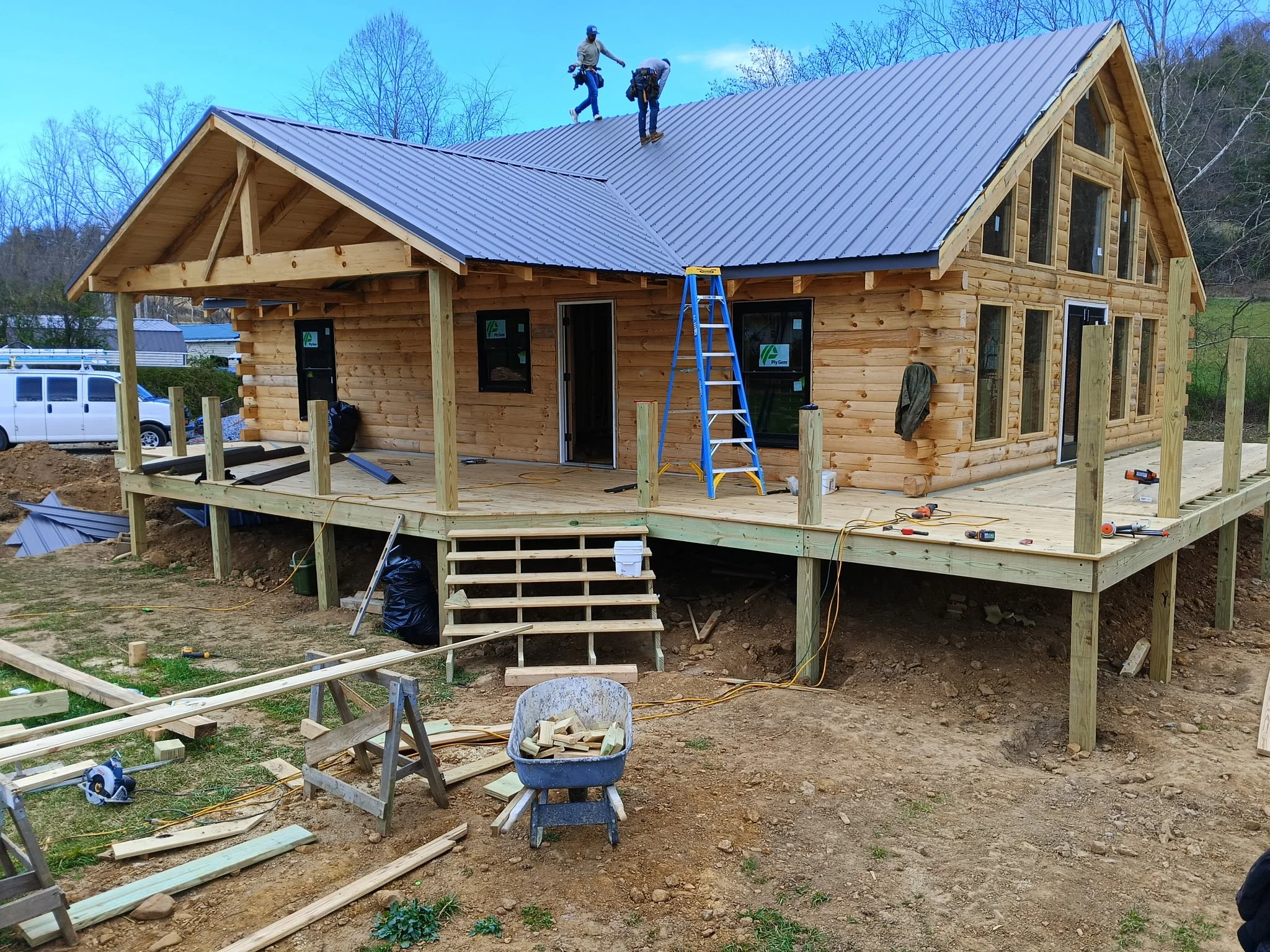 Workers building a wooden house with a deck, metal roof, and multiple windows, on a construction site with tools and materials scattered around.
