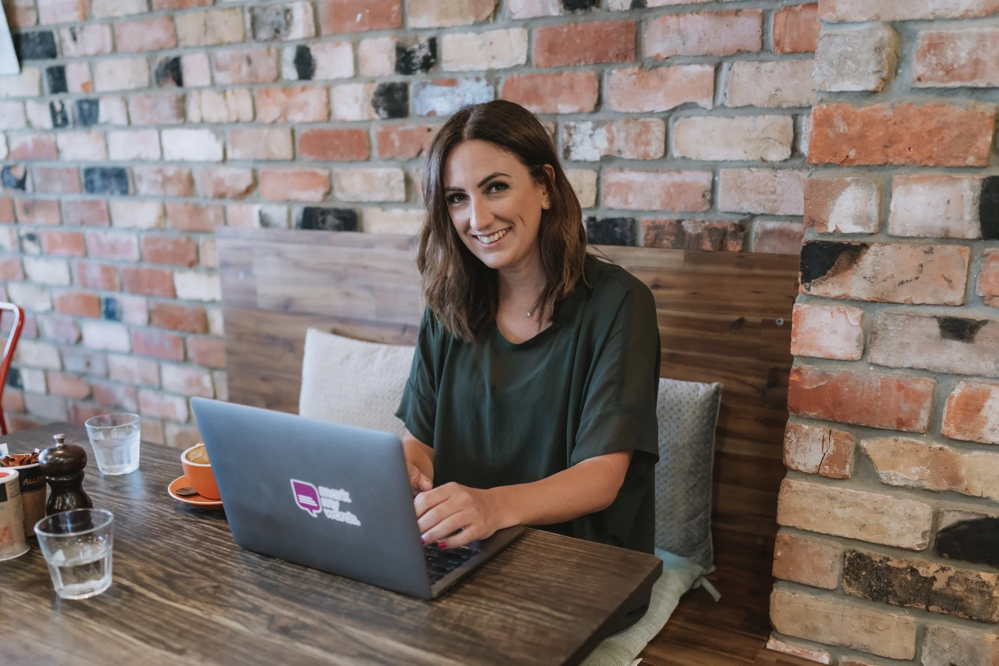 A woman with shoulder-length brown hair smiling while working on her laptop in a cozy cafe setting with an exposed brick wall in the background.