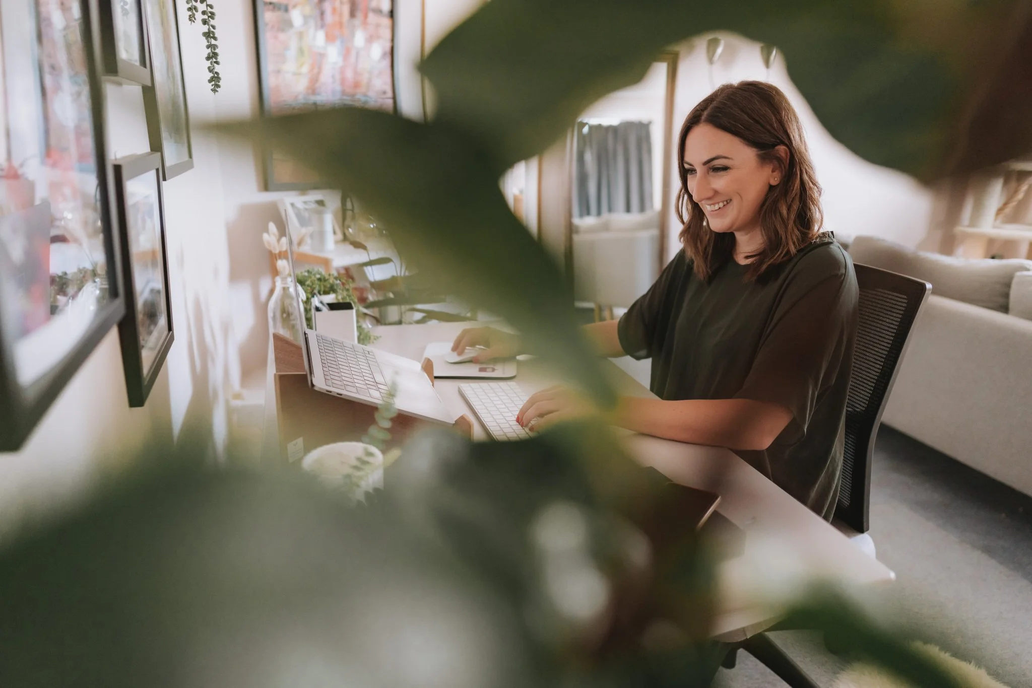 A woman working at her desk with a computer, partially obscured by blurred green leaves in the foreground, in a cozy, well-decorated room.