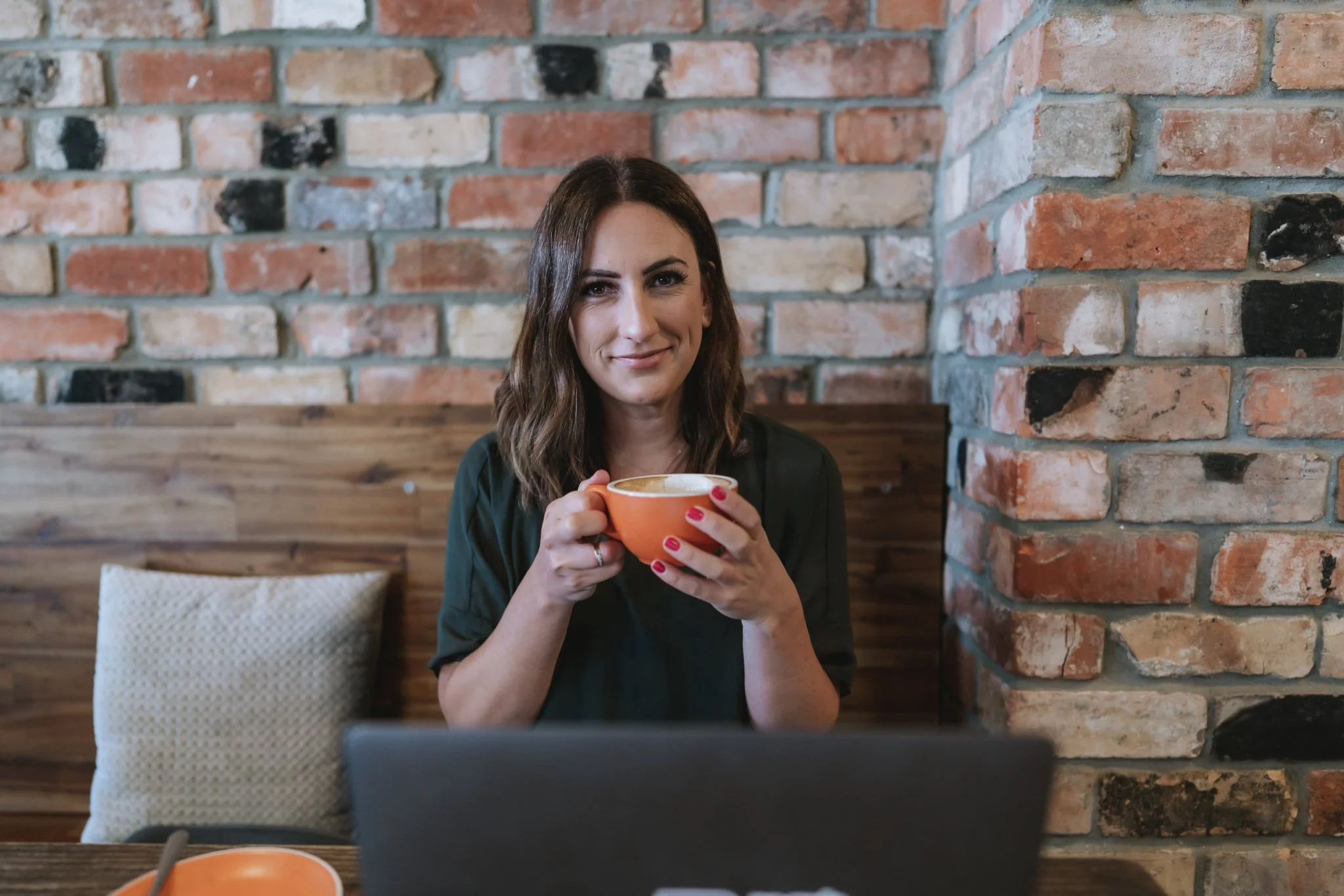 A woman with shoulder-length brown hair sitting at a wooden table in a cafe, holding an orange coffee mug, smiling at the camera. The background features a brick wall and a beige cushion on the bench.