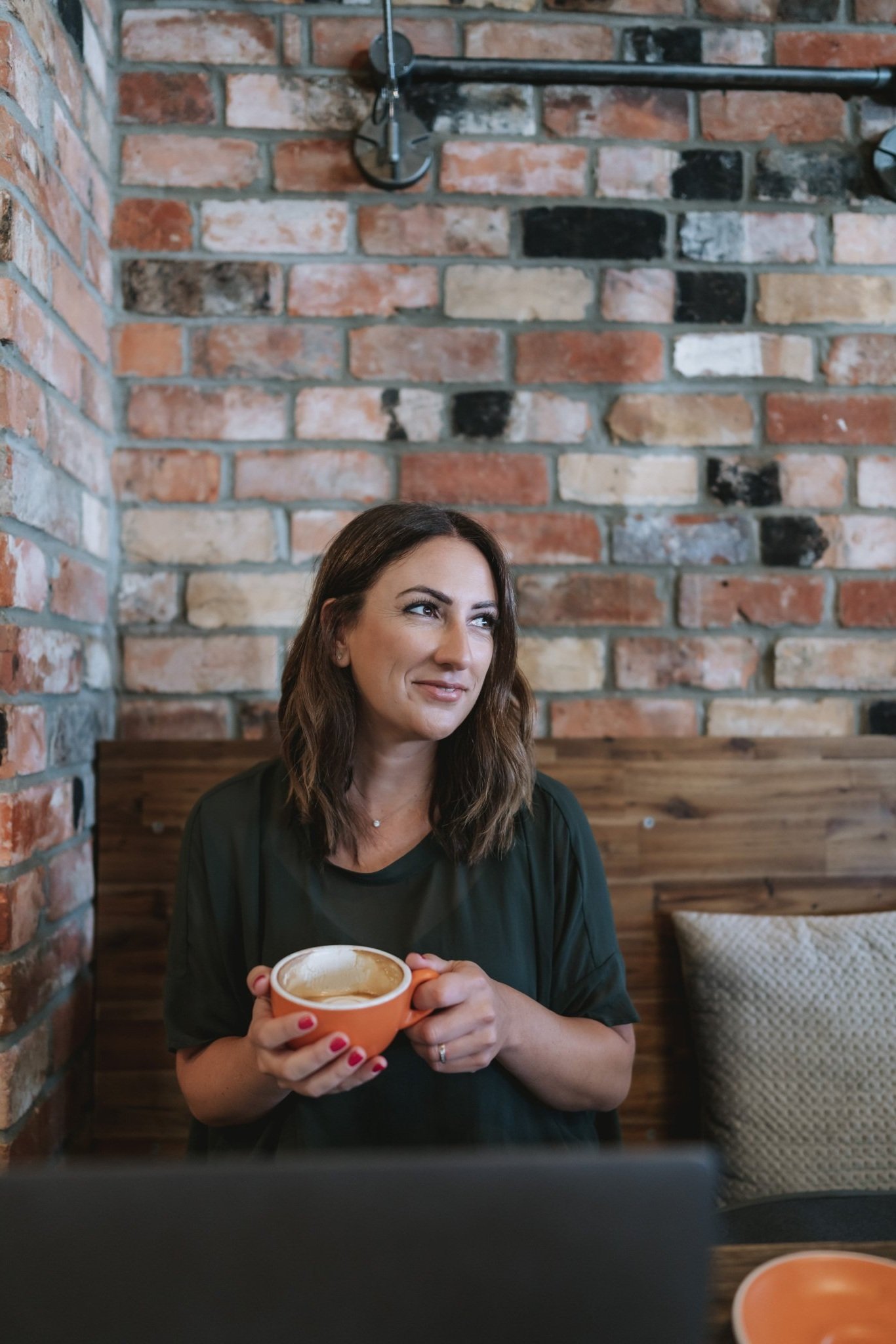 A woman with wavy brown hair holding an empty orange coffee cup, sitting in a cafe with a brick wall background.