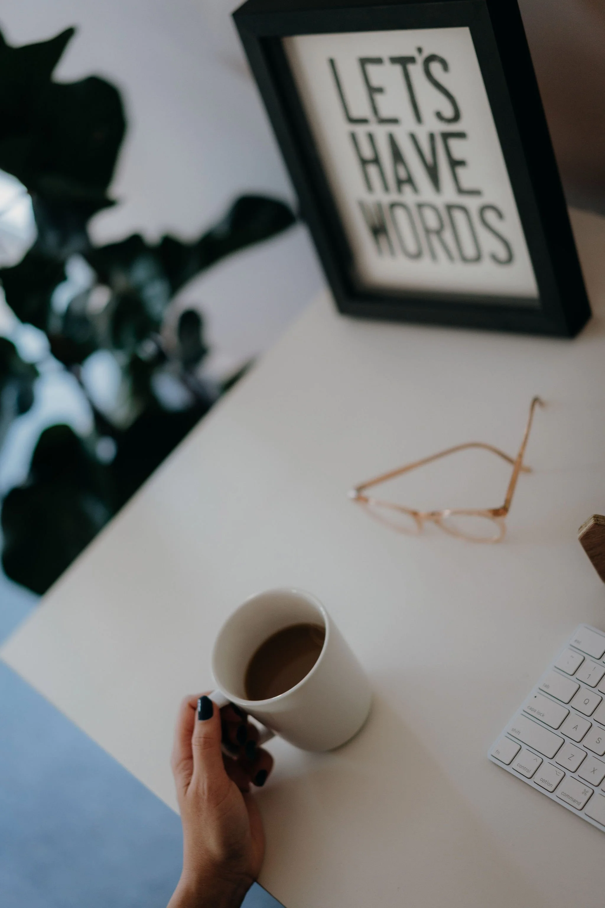 Person holding a white coffee mug at a white desk with a framed sign that reads 'Let's Have Words,' gold-colored glasses, a keyboard, and a potted plant.